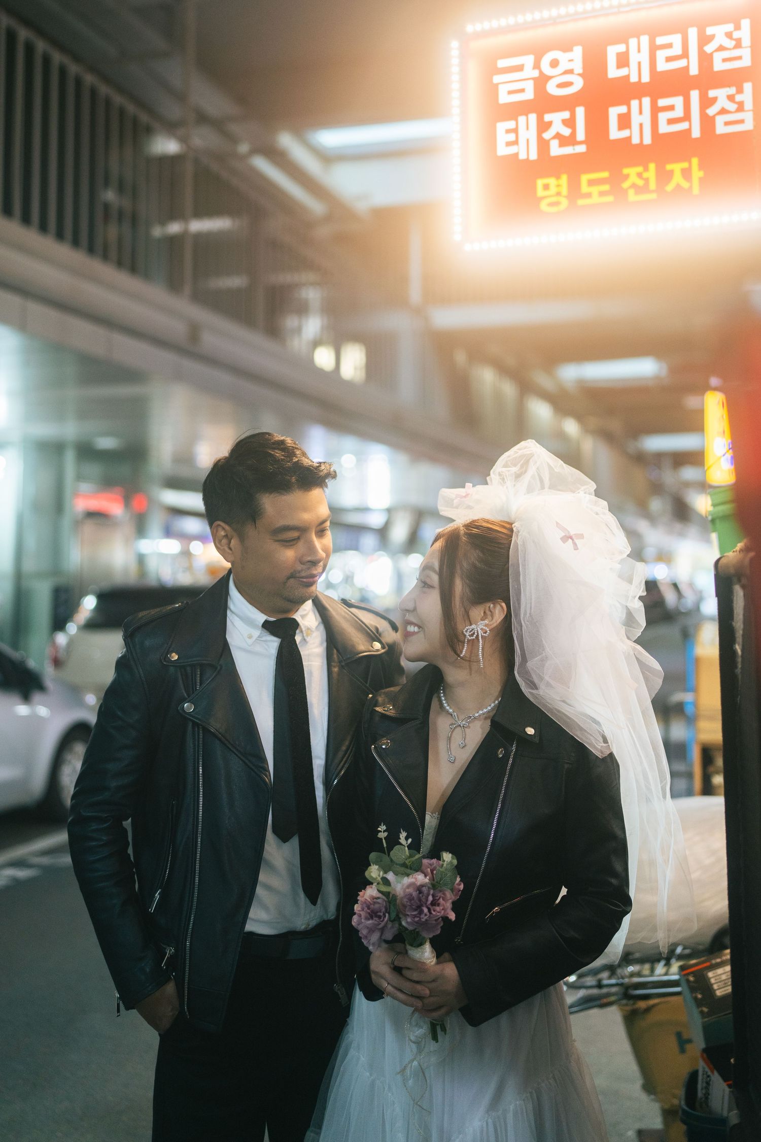 A bride and groom share intimate moments in front of Korean storefronts with neon signs glowing in the background.