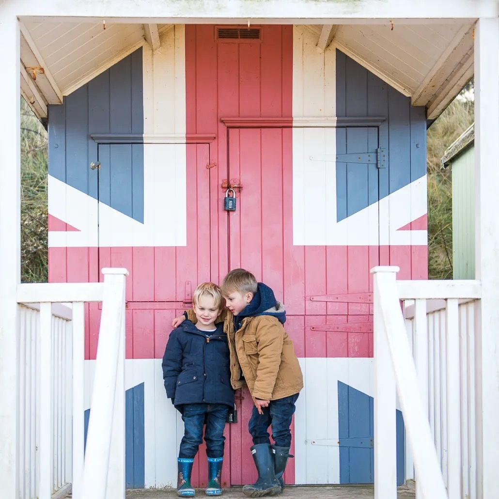 Two children sitting on the steps of a beach hut painted with a red and blue flag design.