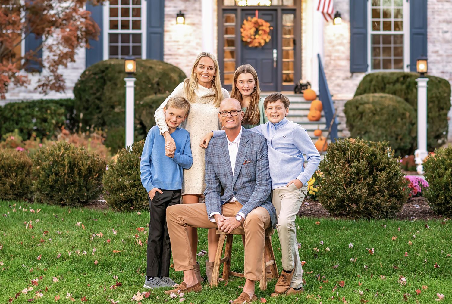 A group sits together on a bench in front of a traditional brick house with autumn decorations and landscaping.