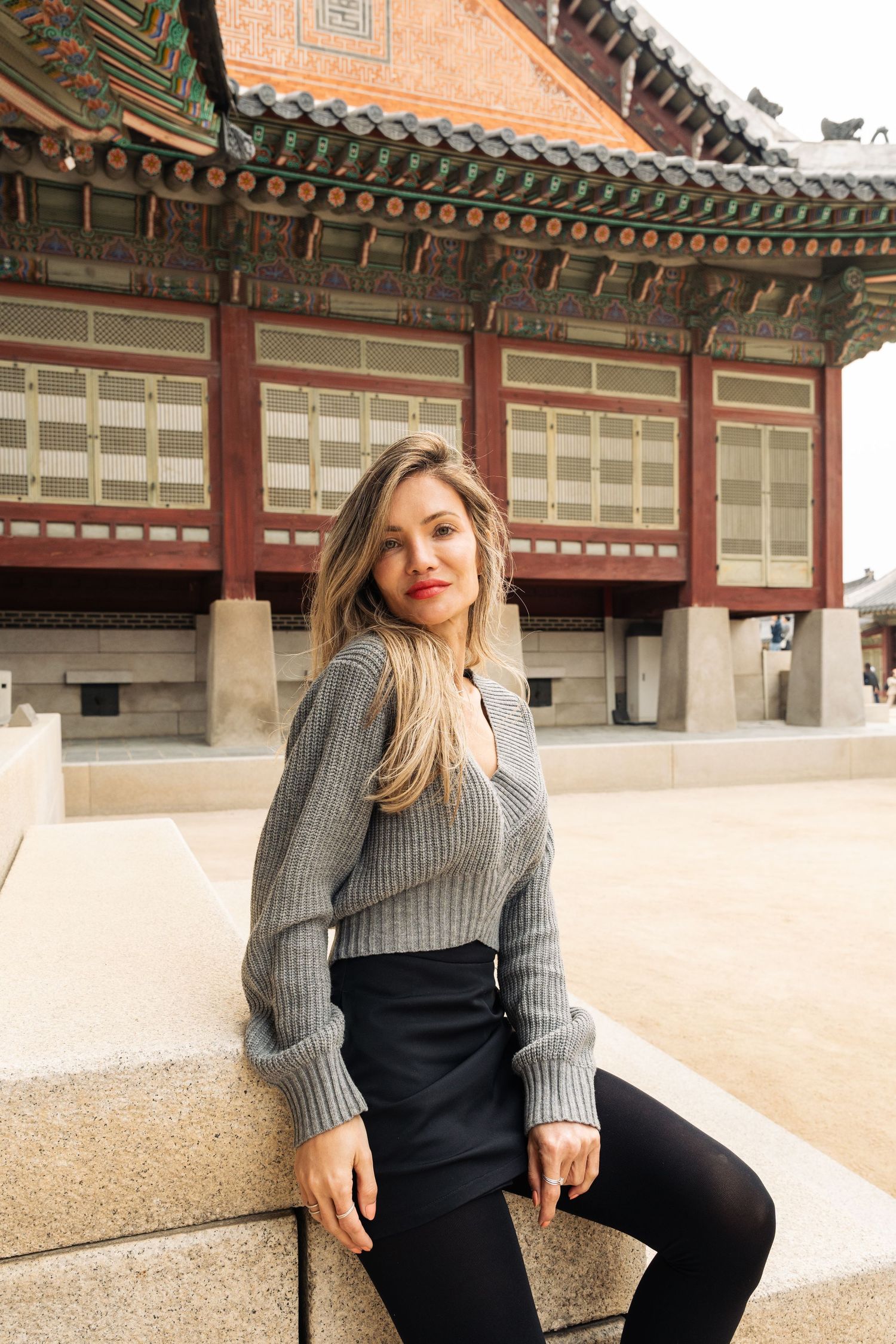 Woman in gray sweater poses at traditional Korean palace courtyard with ornate architectural details and tiled roofs.