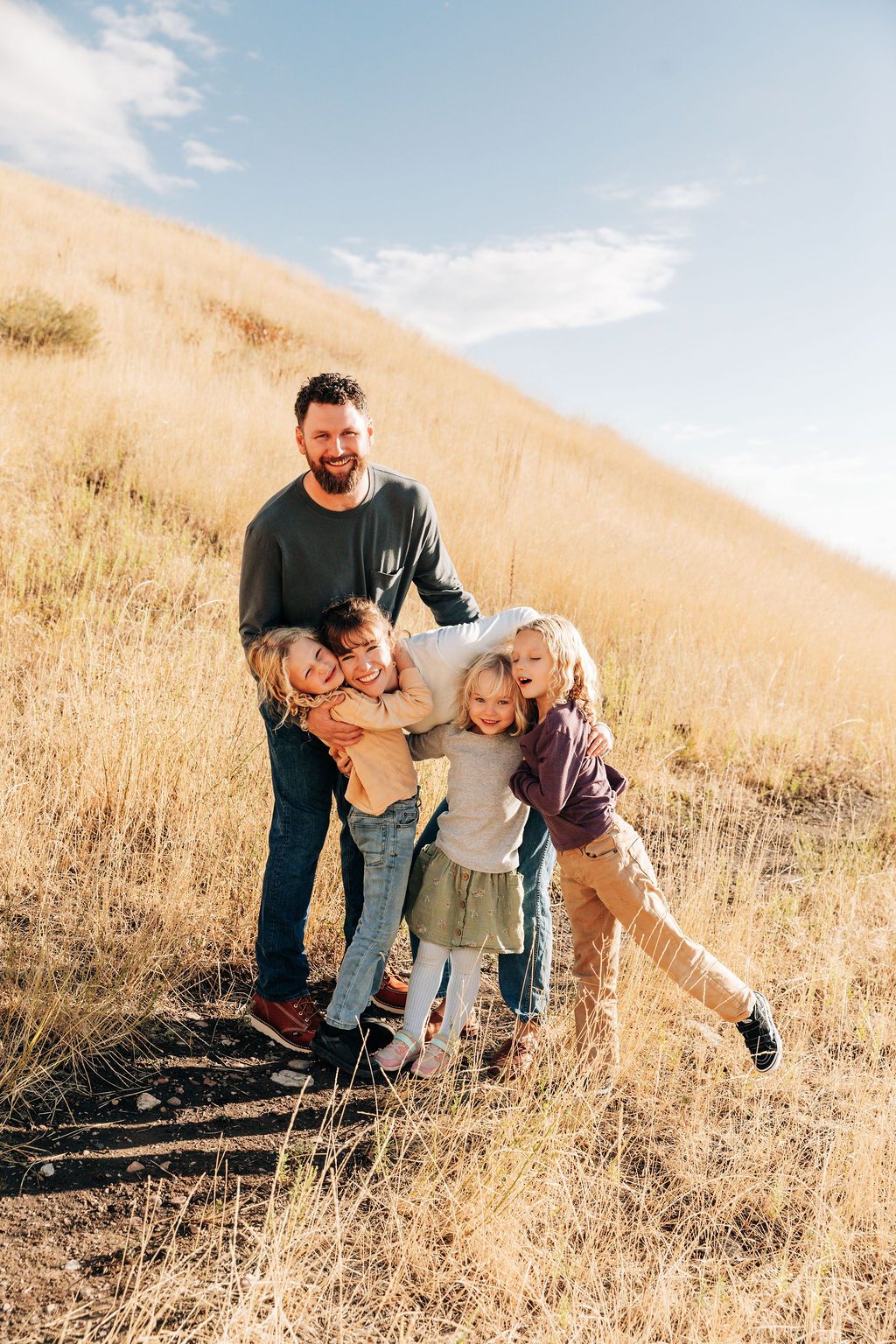 A playful family moment captured in golden sunlit grass on a hillside with joyful expressions and casual attire.