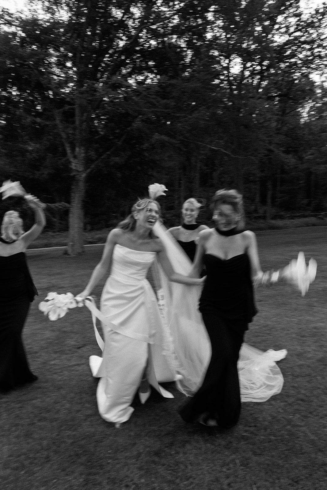 A group in flowing white dresses and black attire dances together in a grassy outdoor setting at dusk.