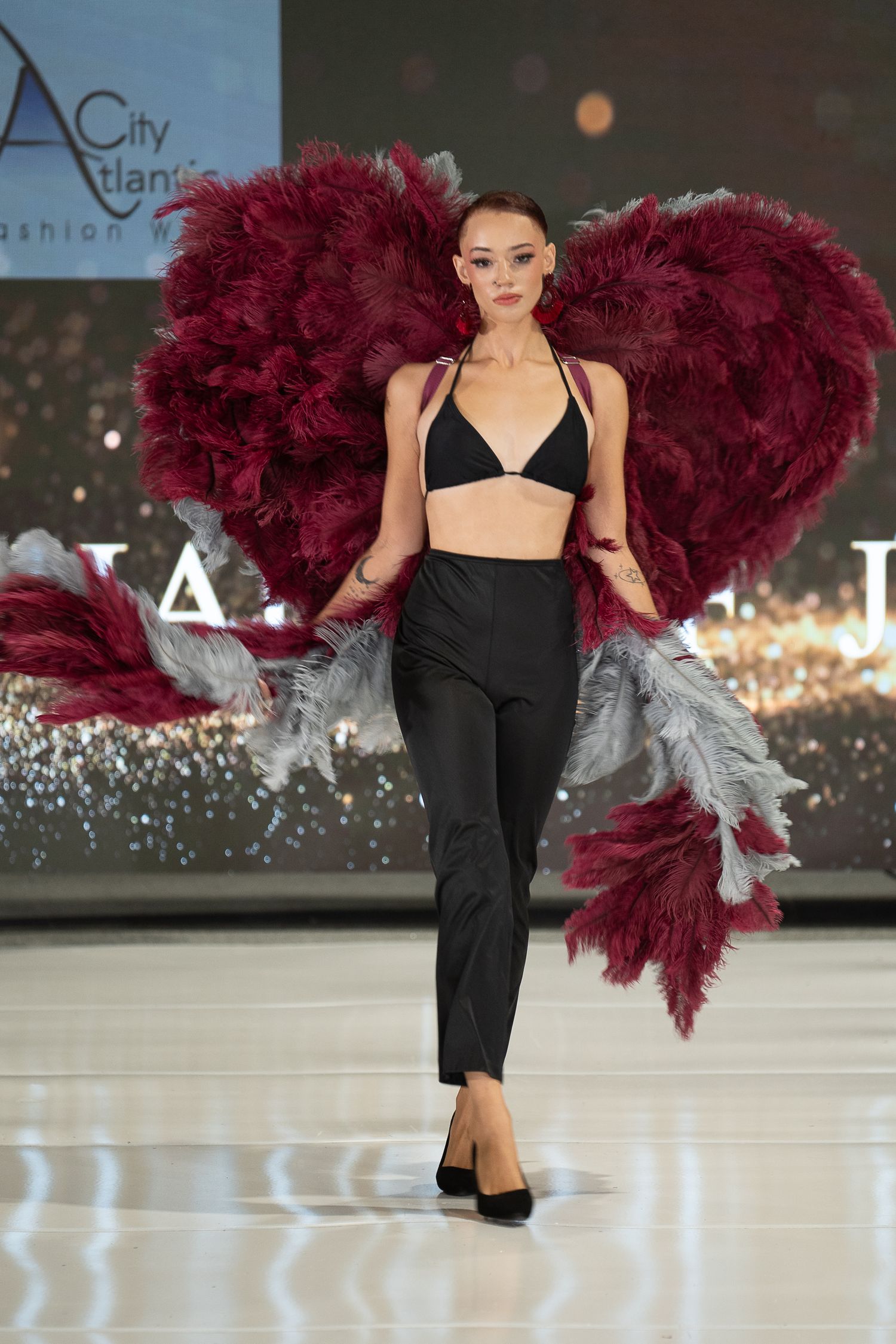 Fashion model walks runway in black jumpsuit with dramatic burgundy feather accessory at fashion show.