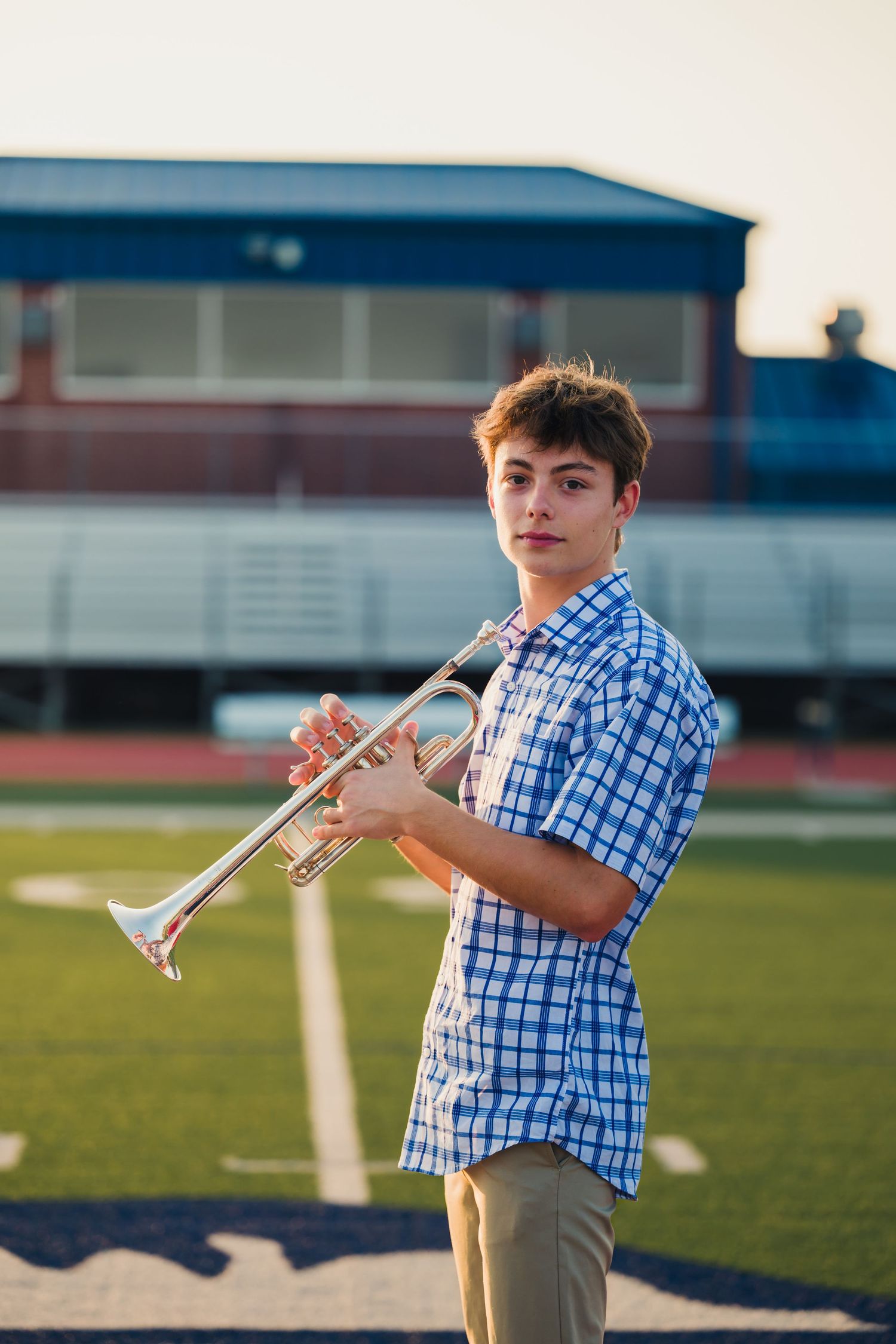Student musician in blue checkered shirt holds trumpet while standing on outdoor field at sunset.