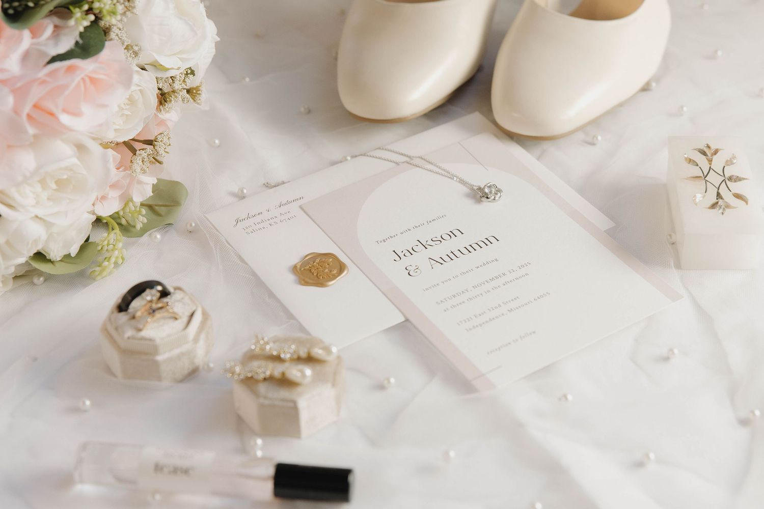 Elegant wedding details including cream shoes, pink roses, invitation card and rings on white linen.