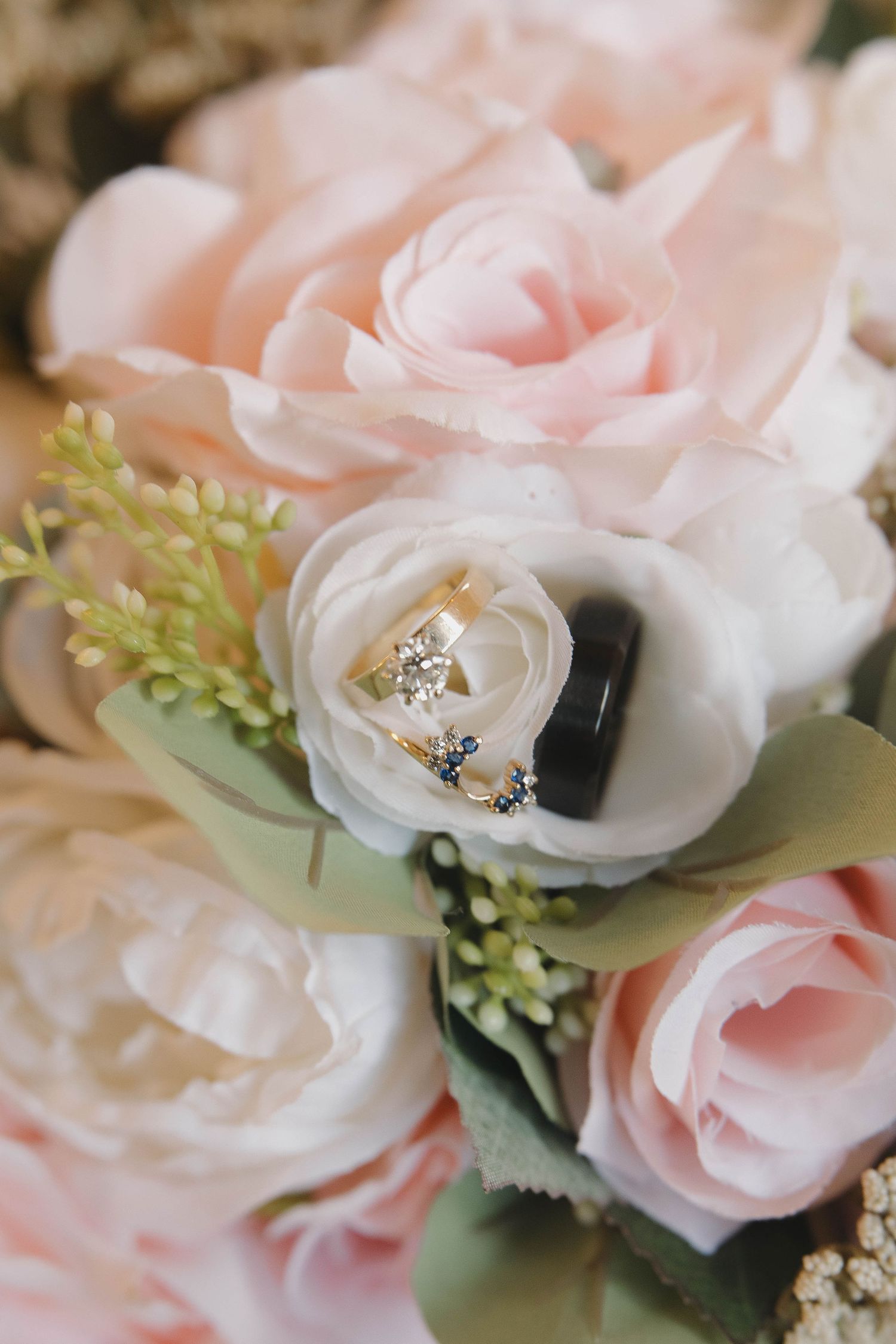 Wedding rings nestled among delicate pink and white roses with green foliage in a romantic bridal bouquet.