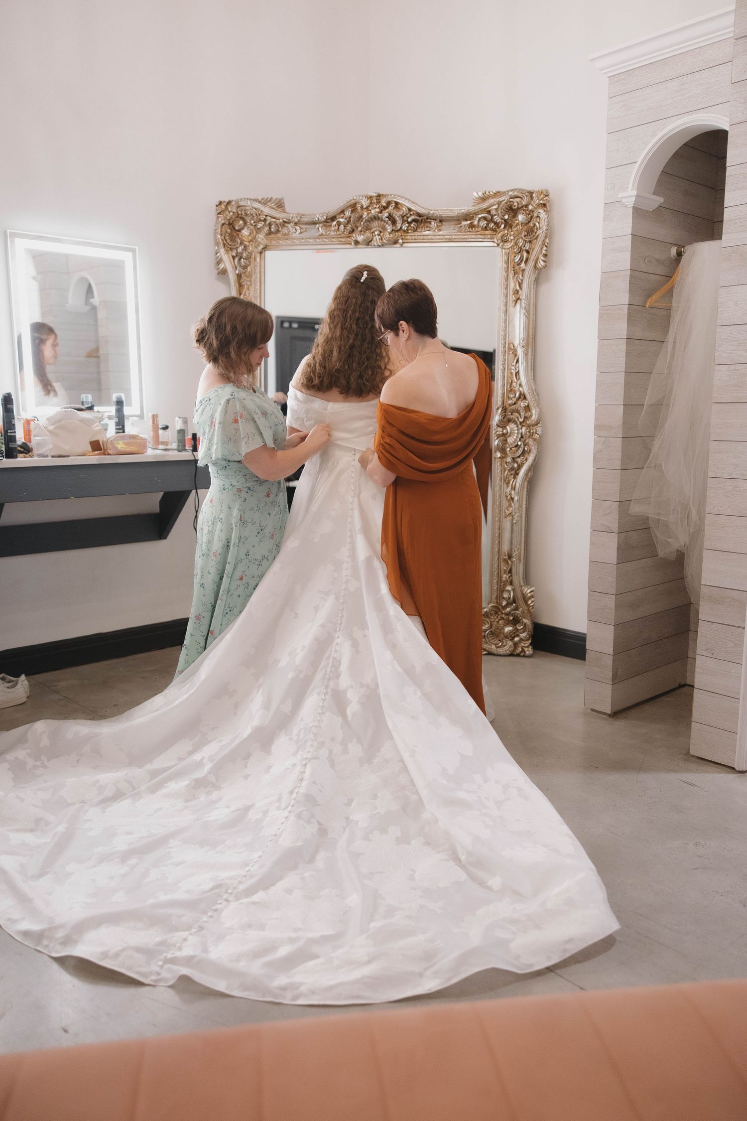A flowing white wedding dress with a long train being adjusted in an elegant white-walled dressing room.