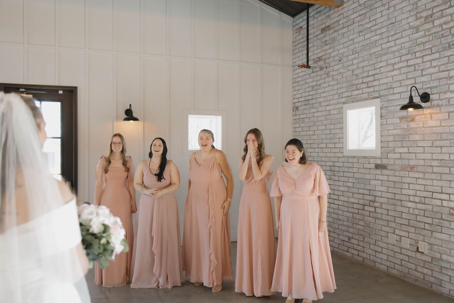 Bridesmaids in blush pink dresses stand together in an industrial-style room with exposed brick walls.
