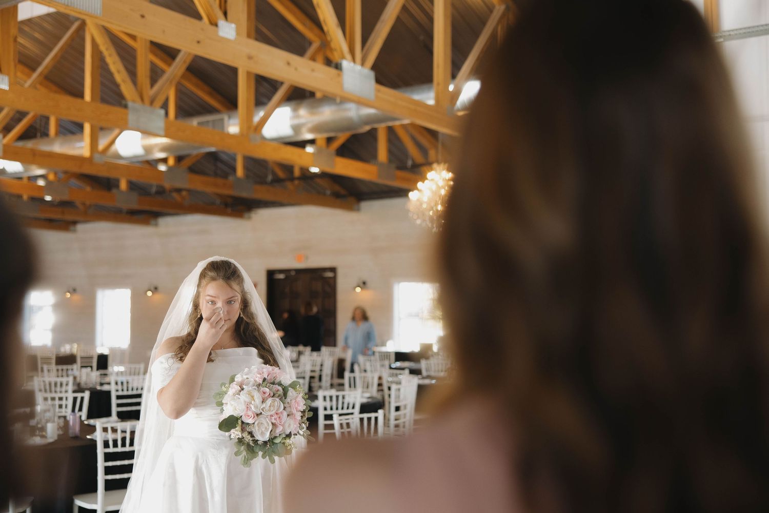 A bride and groom share their first dance in an elegant rustic barn venue with exposed wooden beams and white decor.