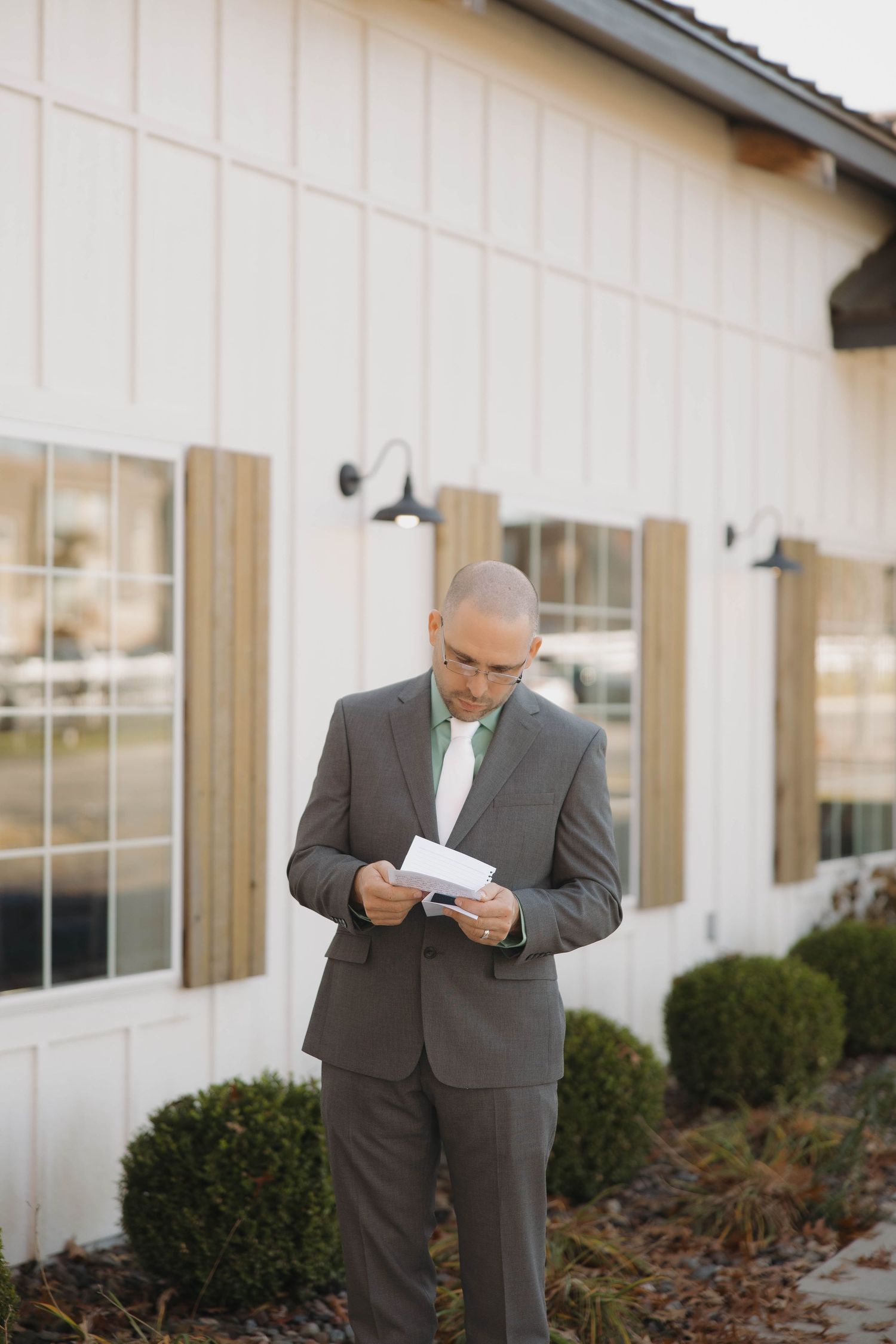 A person in a grey suit reads from papers while standing outside a white building with wooden shutters.
