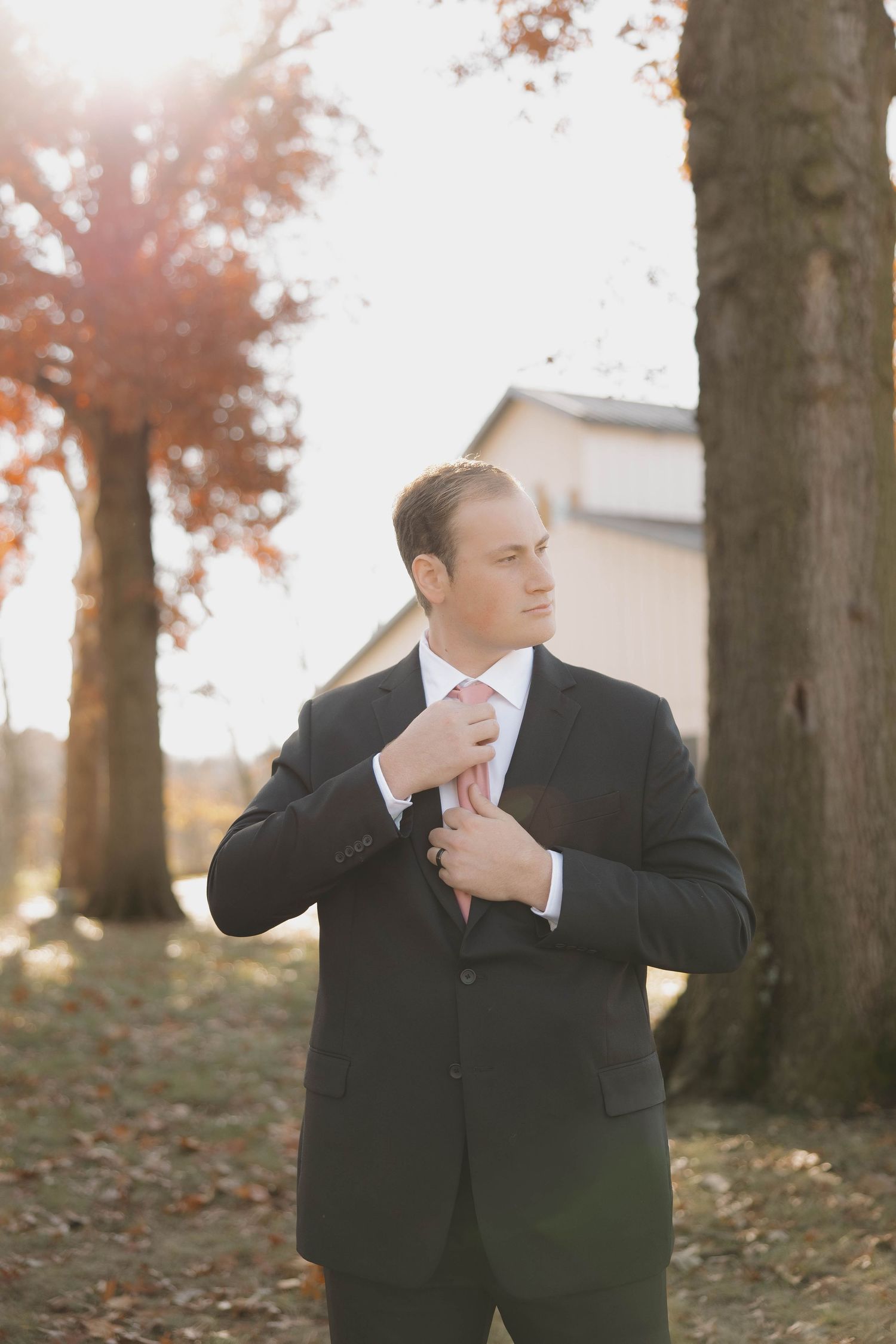 A person in a black suit adjusts their tie while standing among autumn trees.