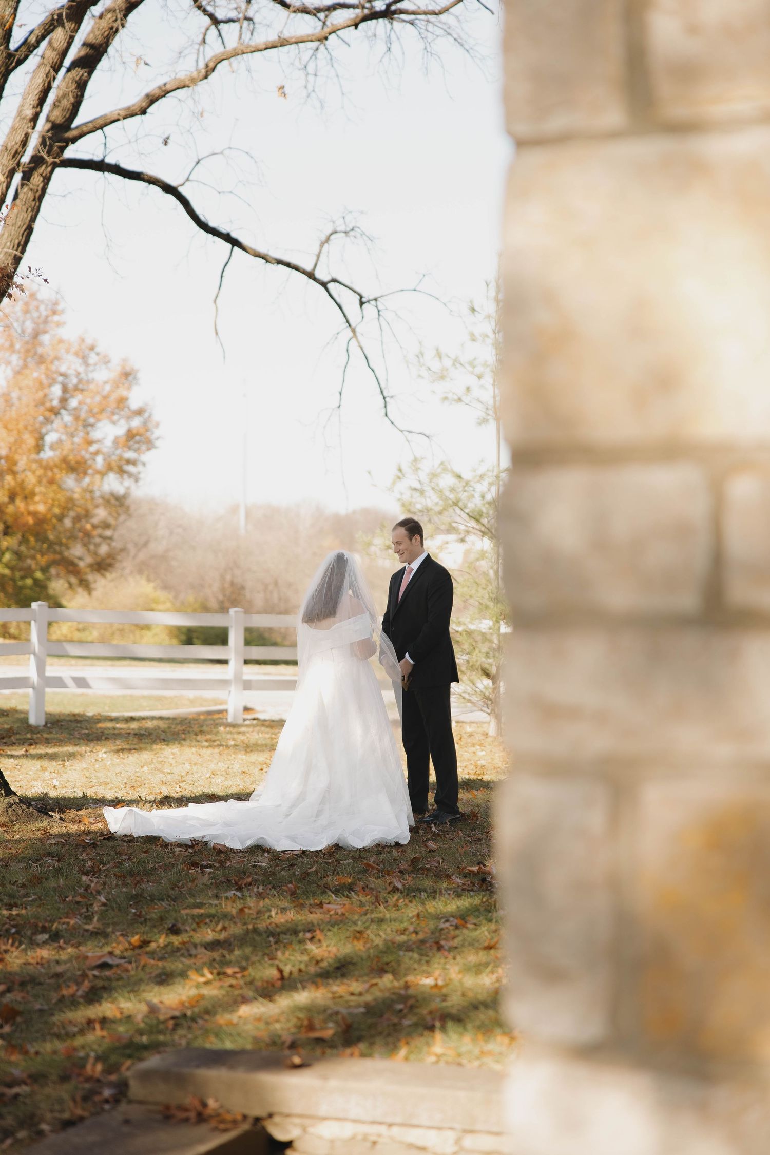 A couple shares an intimate moment in their wedding attire near a white fence and autumn trees.
