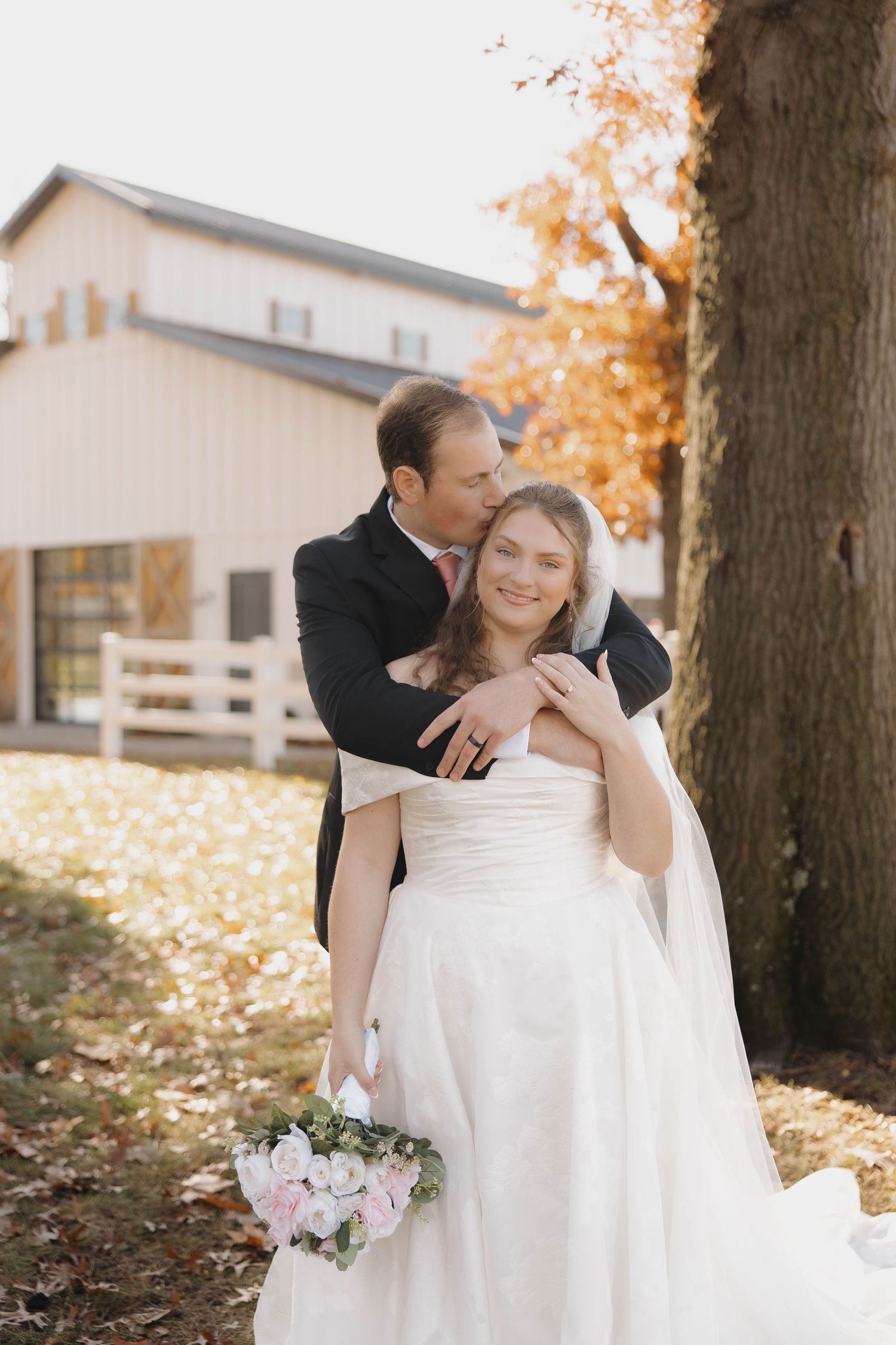 A romantic autumn wedding photo of a couple embracing by a rustic barn with falling leaves.