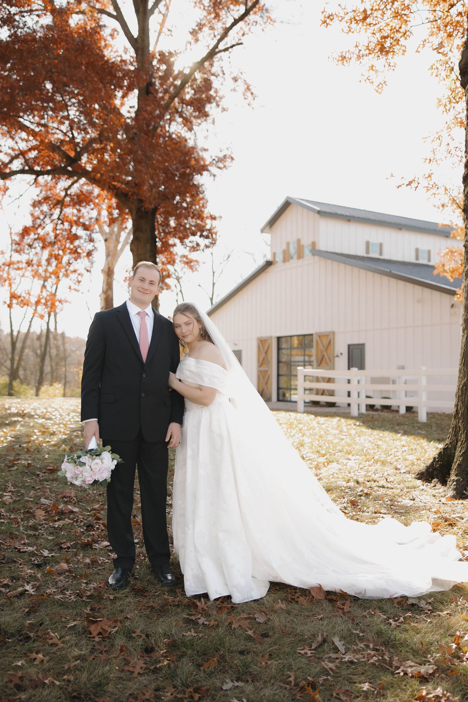 A wedding couple poses in front of a rustic white barn during autumn with orange fall foliage in the background.