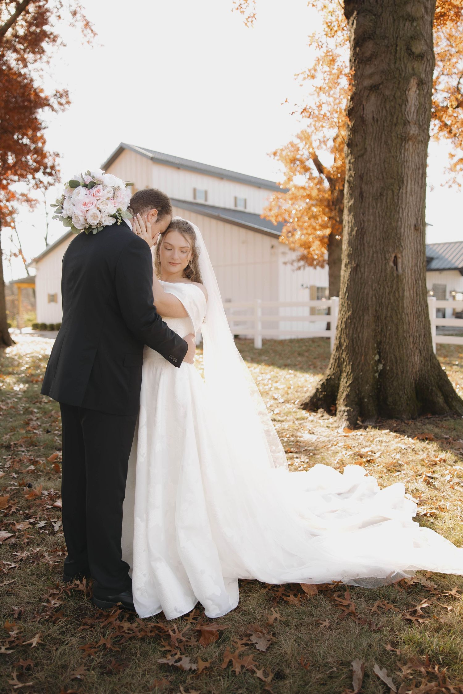 A couple in wedding attire embraces under fall foliage with a white building in the background.
