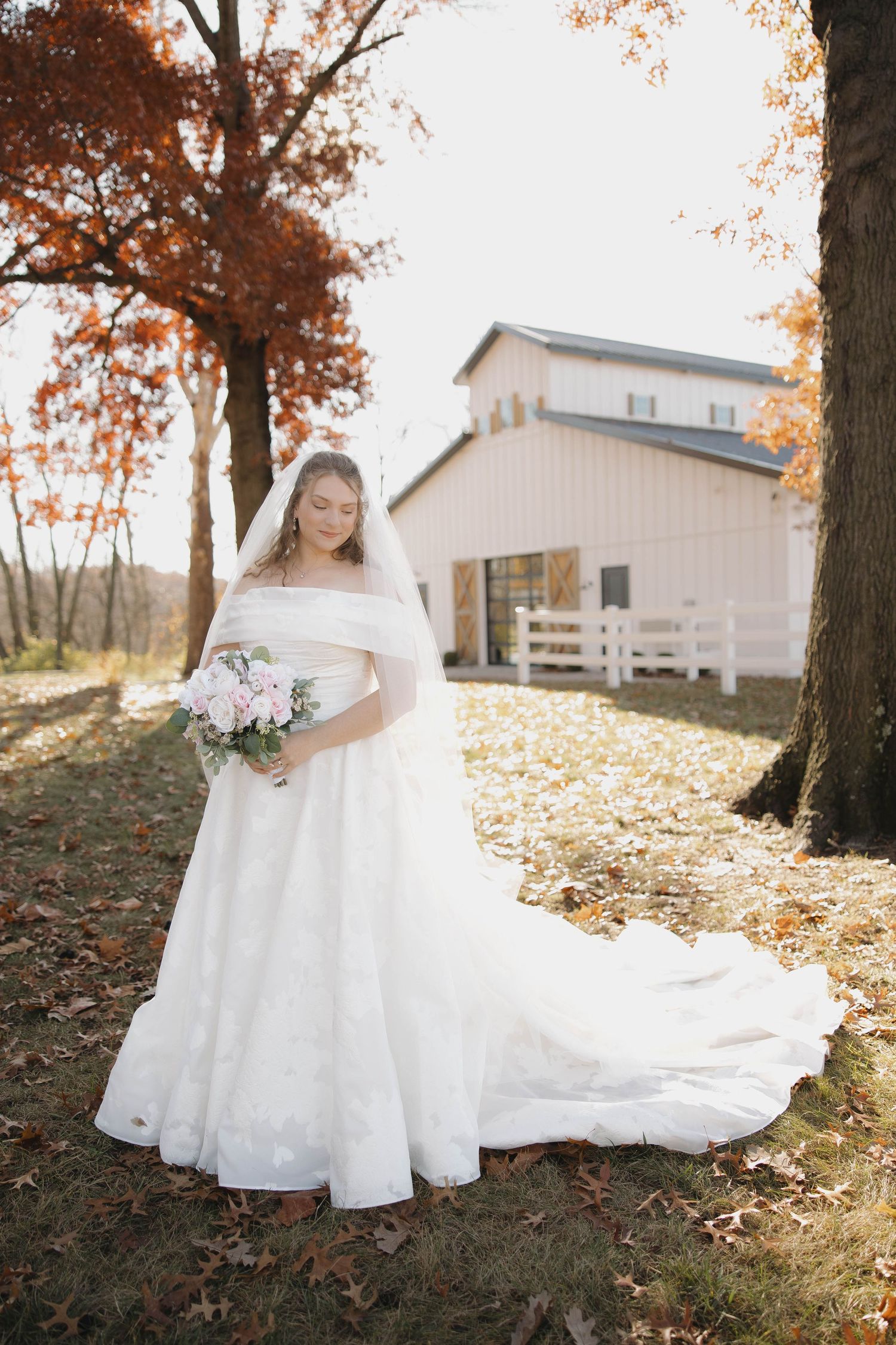 A white wedding dress flows elegantly across fallen autumn leaves on a hillside with white barns in the background.