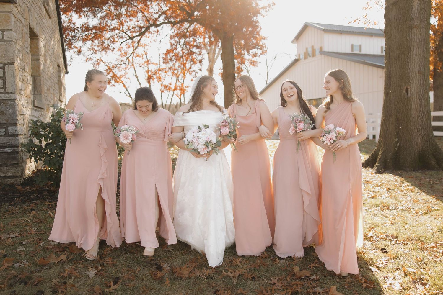 A bridal party in blush pink dresses poses together outdoors on an autumn day with golden sunlight.