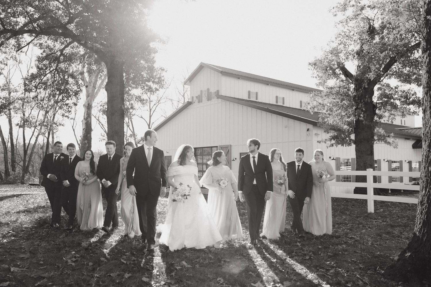 Wedding party poses in front of white barn on autumn day in black and white photograph.