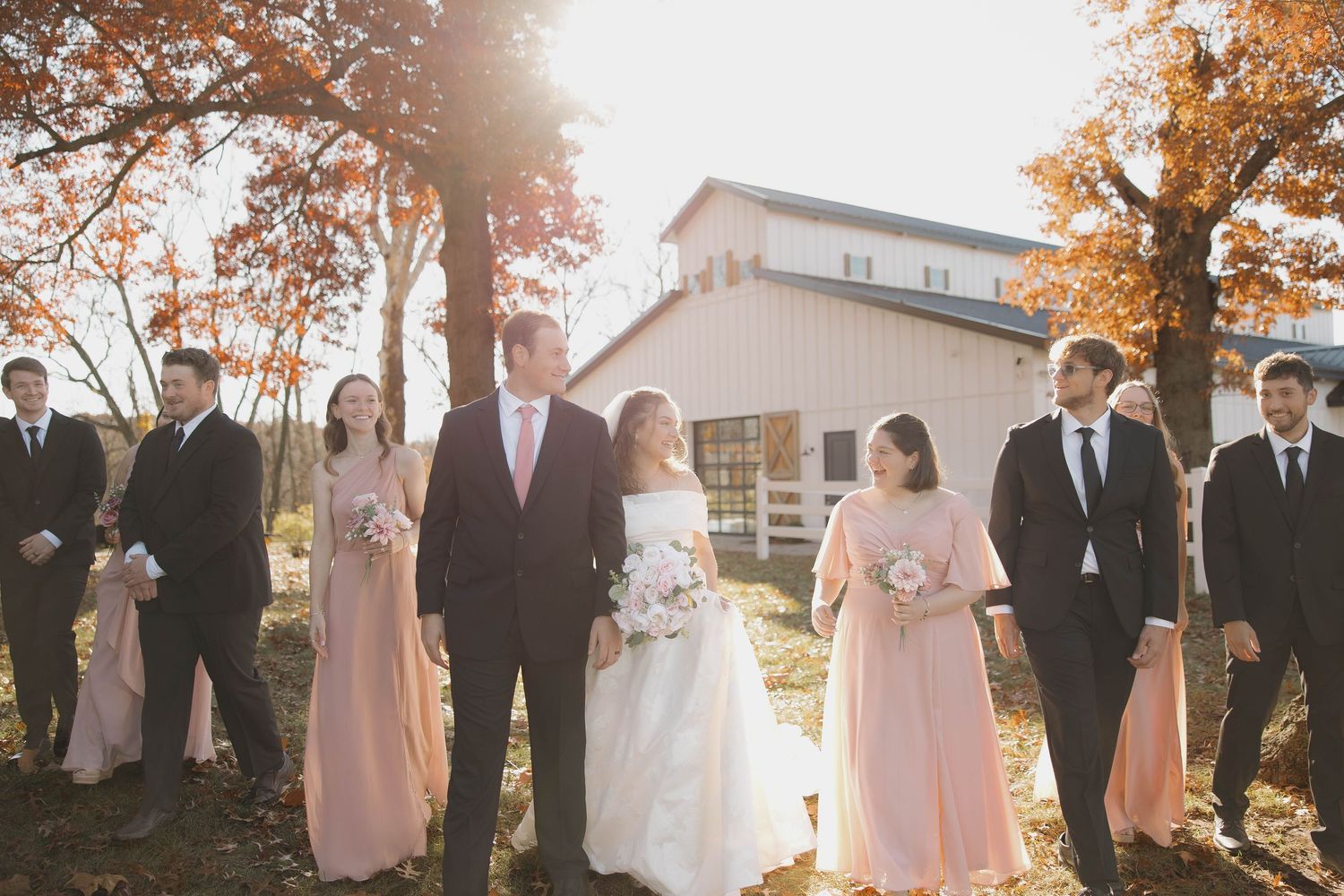 Wedding party stands together under autumn trees with golden sunlight streaming through the branches.