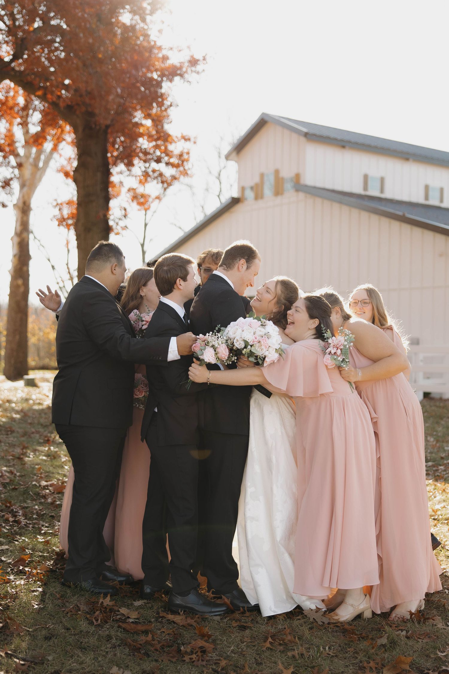 Wedding party in pink dresses and black suits gather for a group photo outside a white barn on an autumn day.
