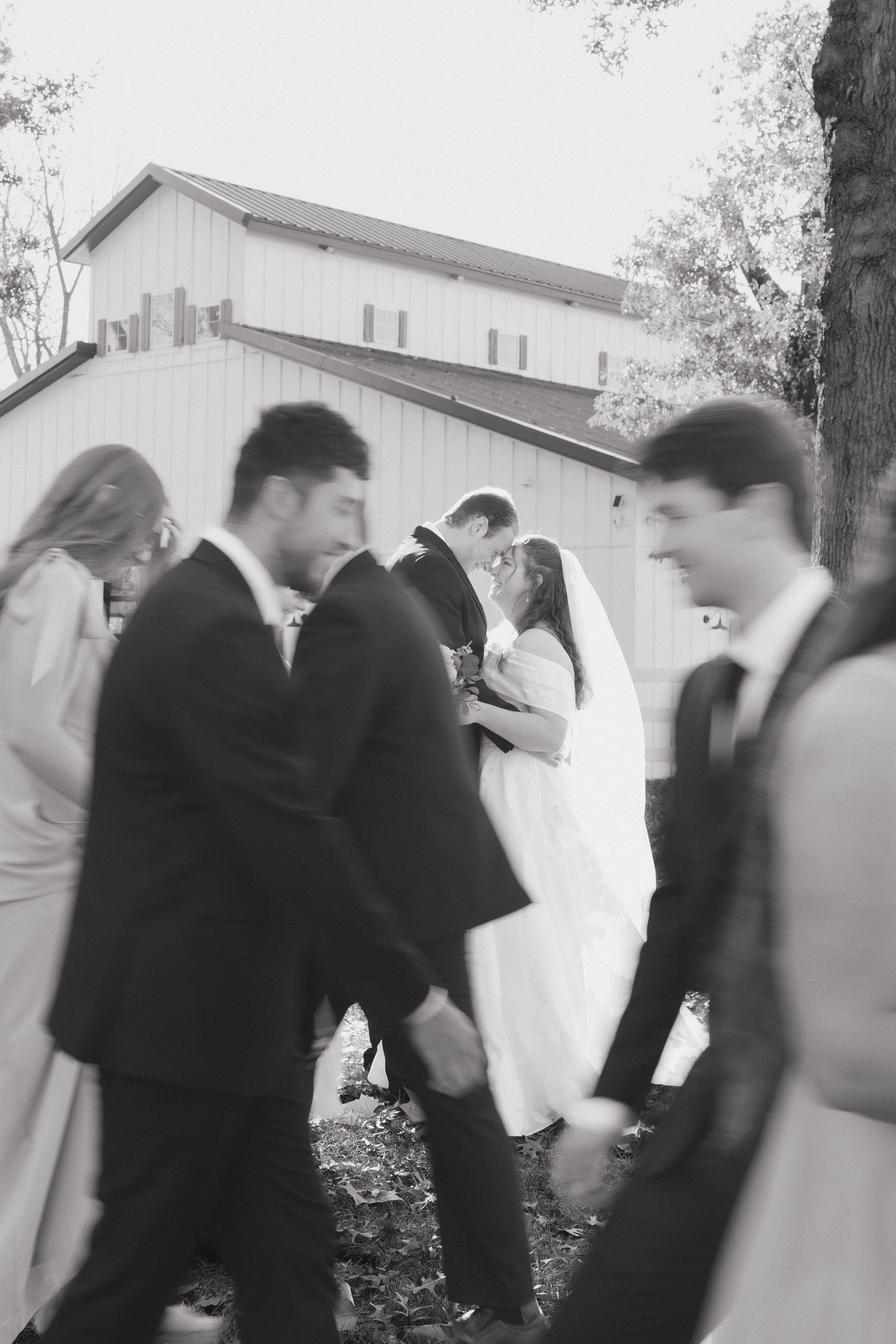 A black and white motion-blurred photo of a wedding party gathered outside a barn.