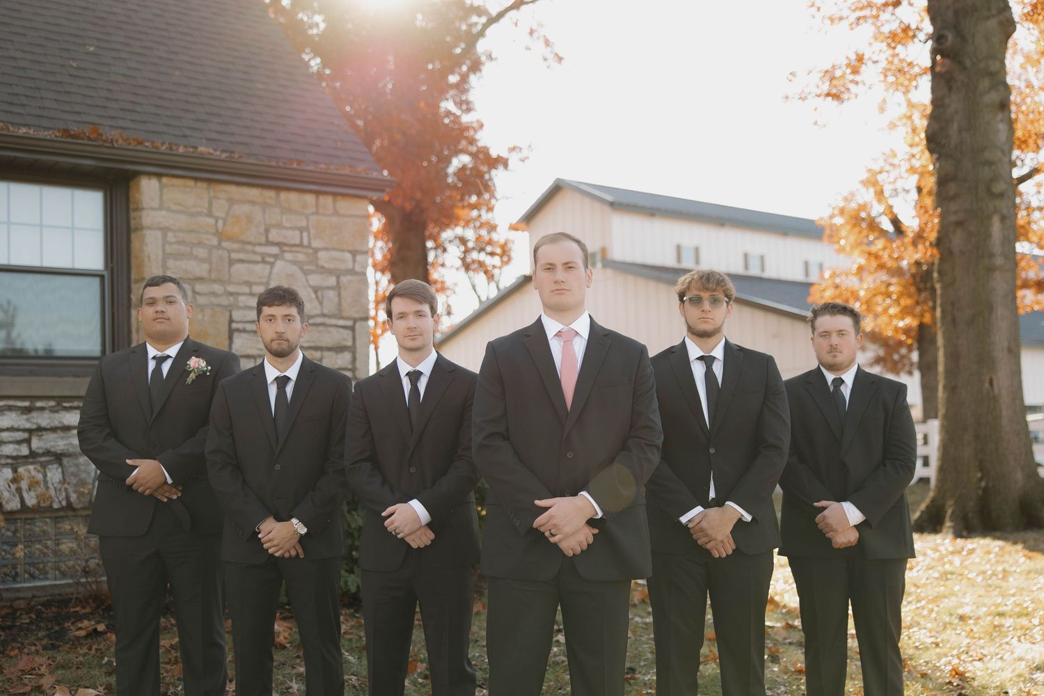 Wedding party in black suits poses outdoors during fall season.