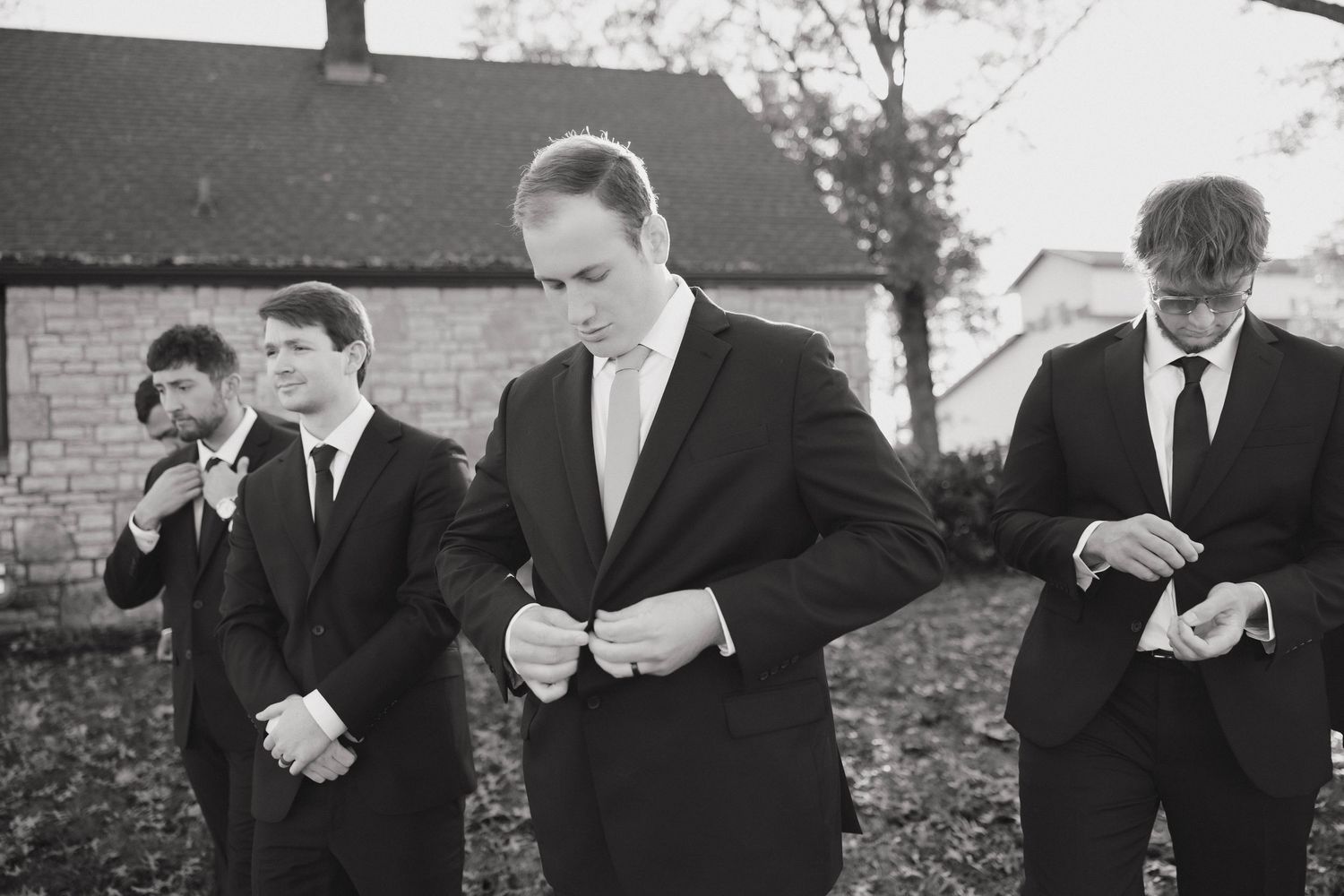 Groomsmen in black suits and ties adjusting their attire outside on a fall day.
