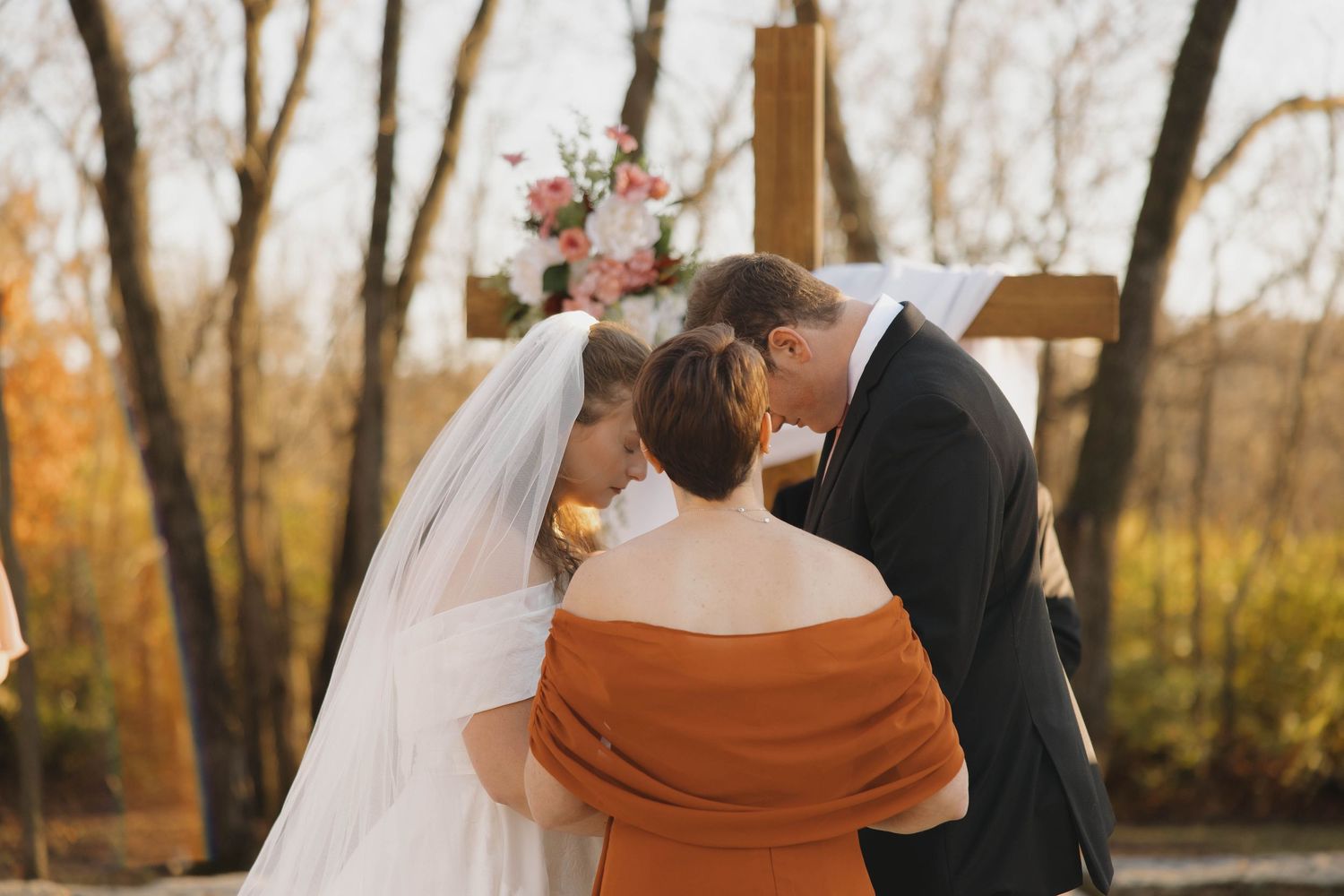 Couple exchanges vows under wooden cross with autumn trees in background.