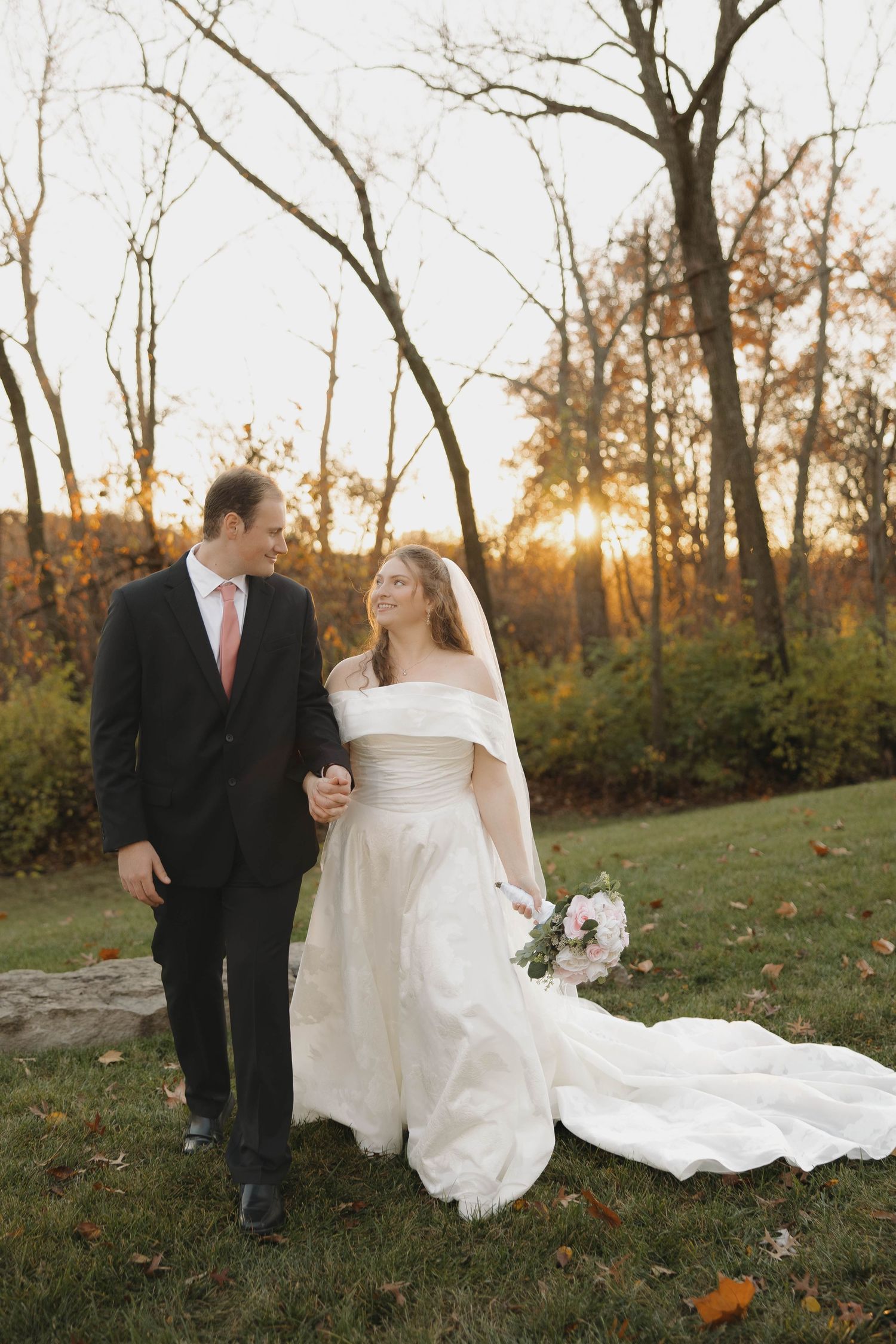 Newlyweds walk hand in hand during sunset with autumn foliage in background.