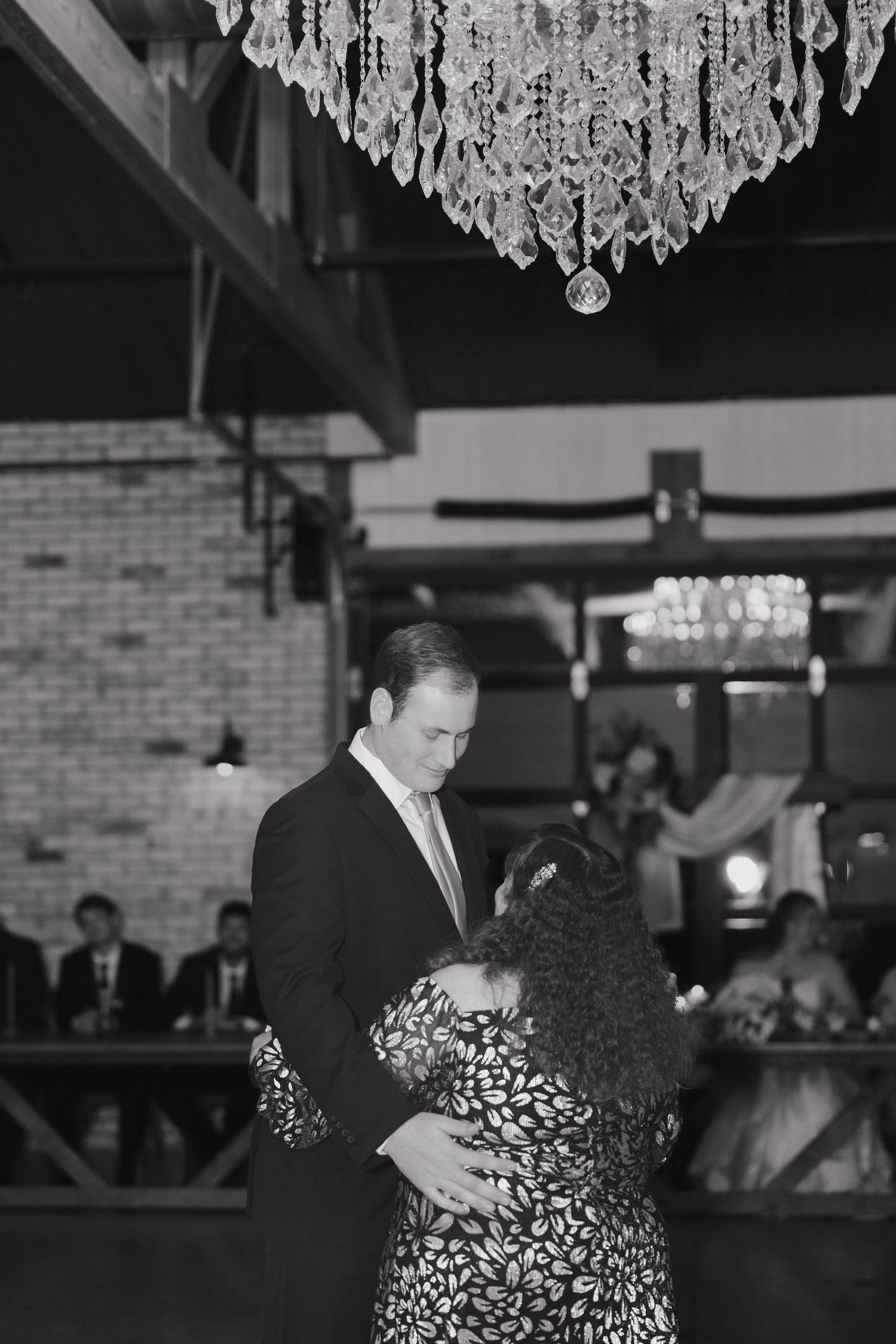 A couple dances beneath an elegant crystal chandelier in a rustic brick-walled venue.