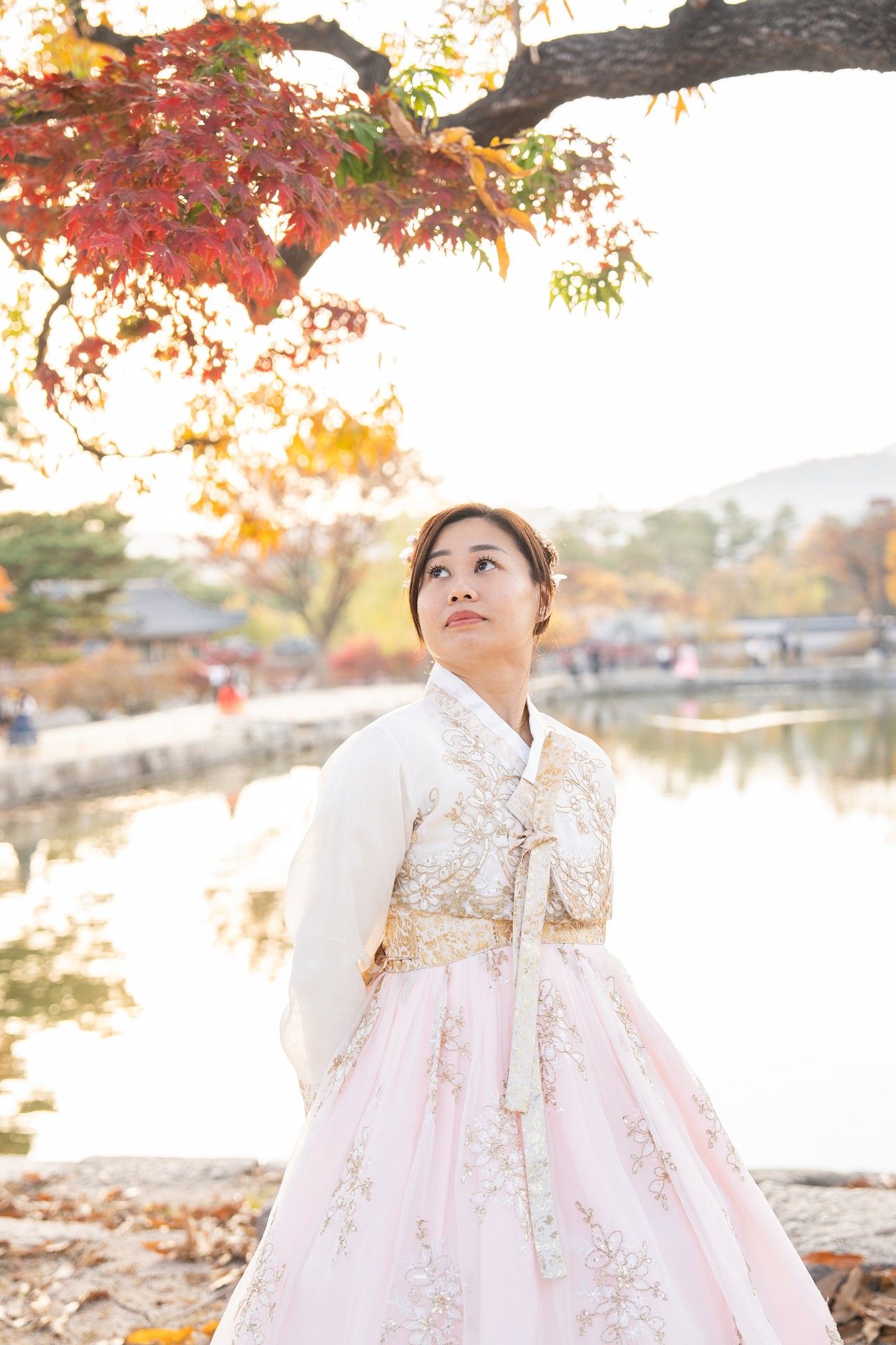 Traditional Korean hanbok dress shown in autumn setting with red maple leaves and lake view in background.