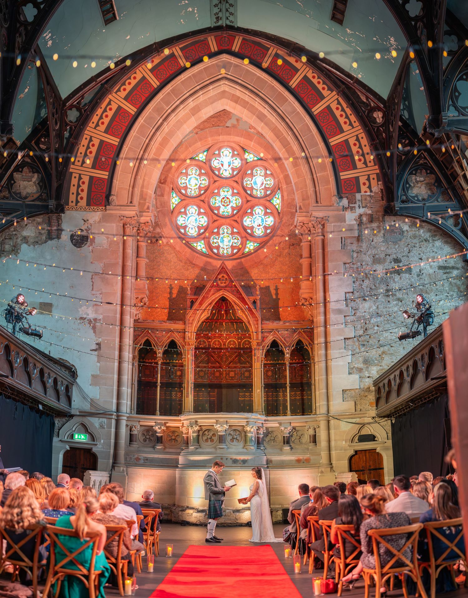 A historic church venue decorated with string lights and cross-back chairs for an elegant wedding ceremony setup. Cottiers.
