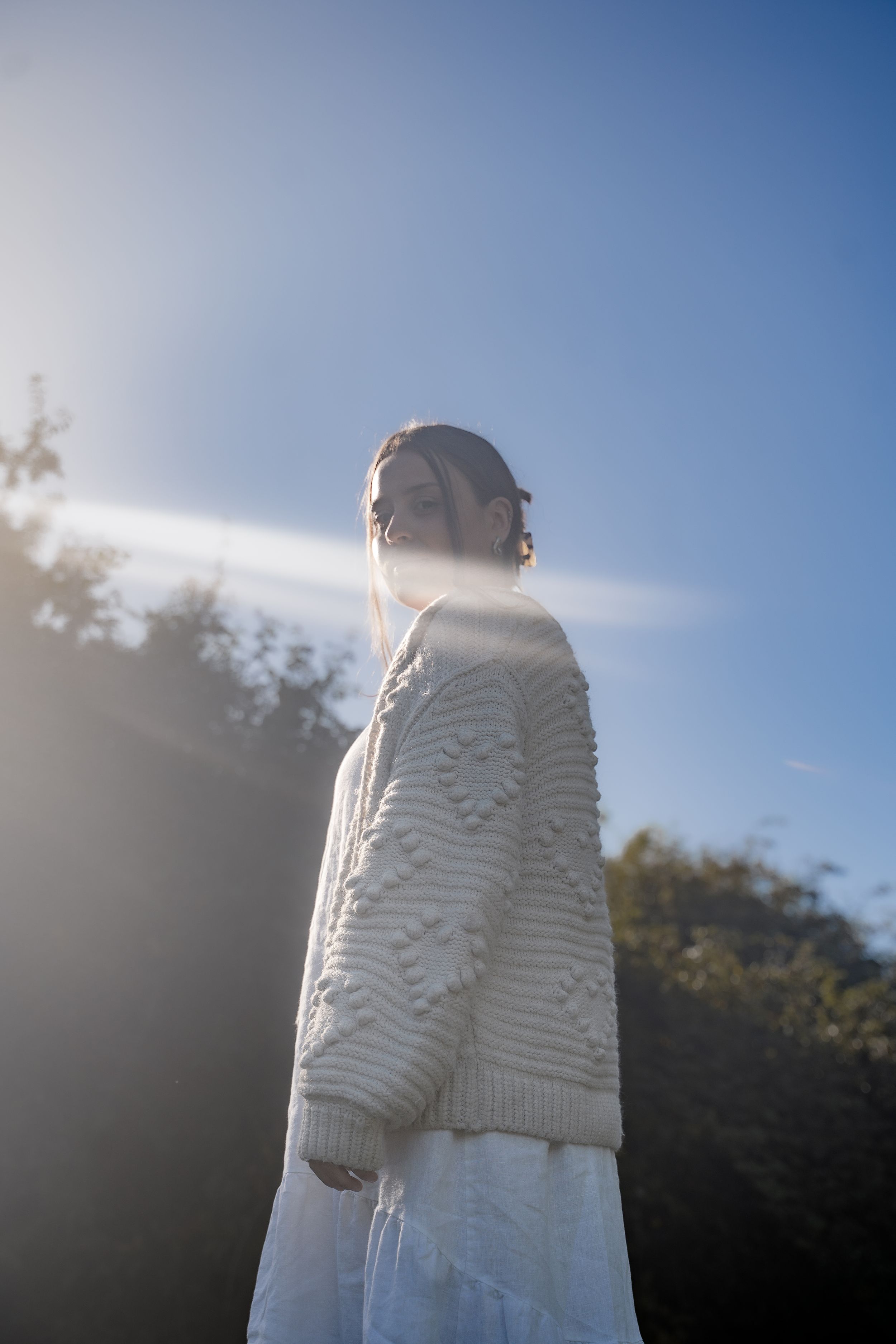 A person wearing a white knit sweater stands against a bright blue sky with sunlight creating a backlit silhouette effect.