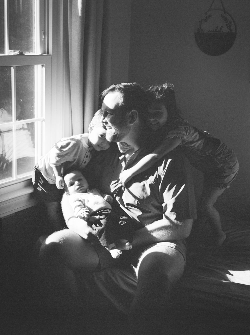 Black and white series showing intimate family moments by a window.