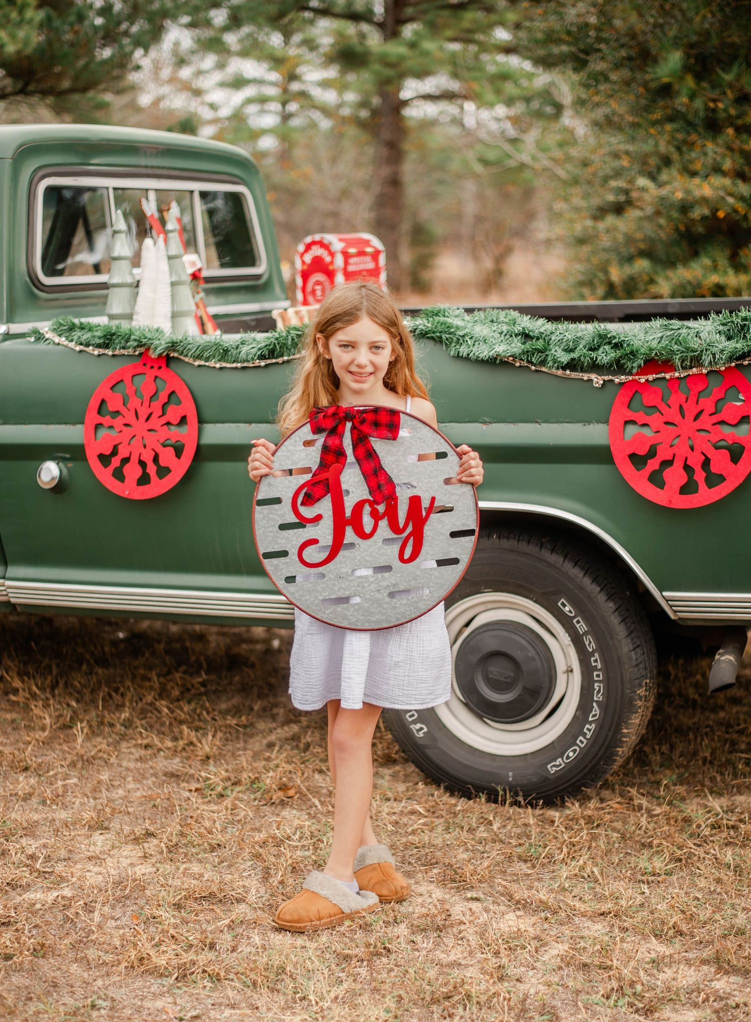 A vintage green truck decorated with red holiday ornaments and a 'Joy' sign creates a festive Christmas scene outdoors.