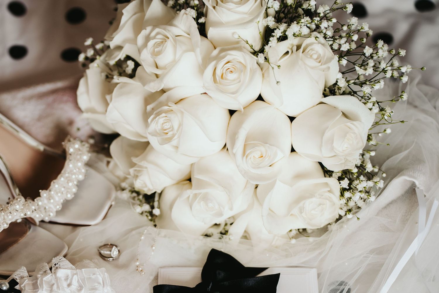 Elegant white rose and baby's breath wedding bouquet with scattered diamond accents on light fabric background.