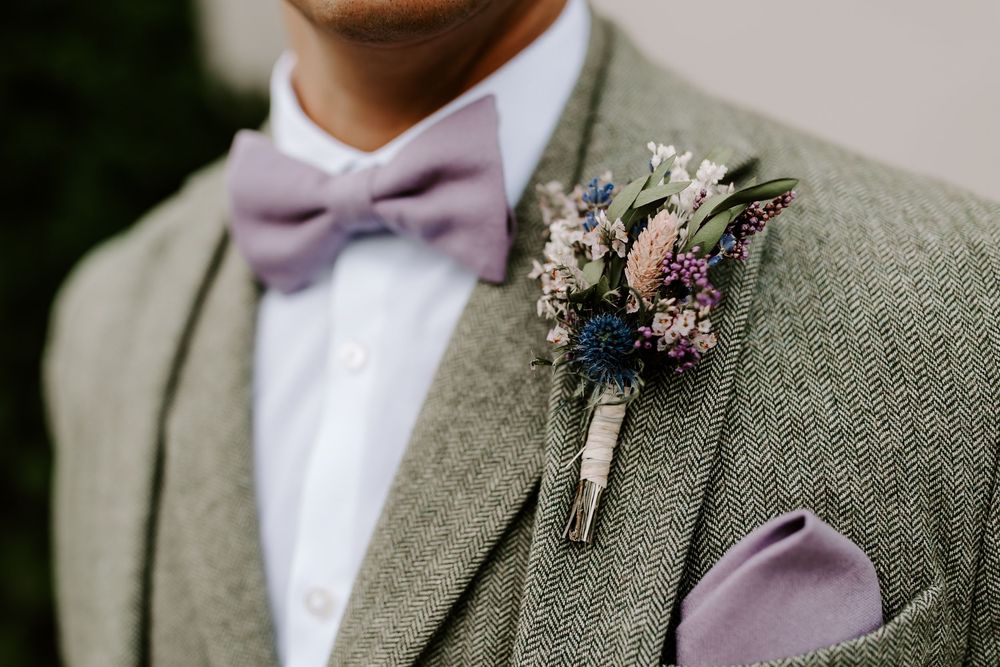 Close-up of sage green suit jacket with lavender bow tie and dried floral boutonniere in rustic wedding style.
