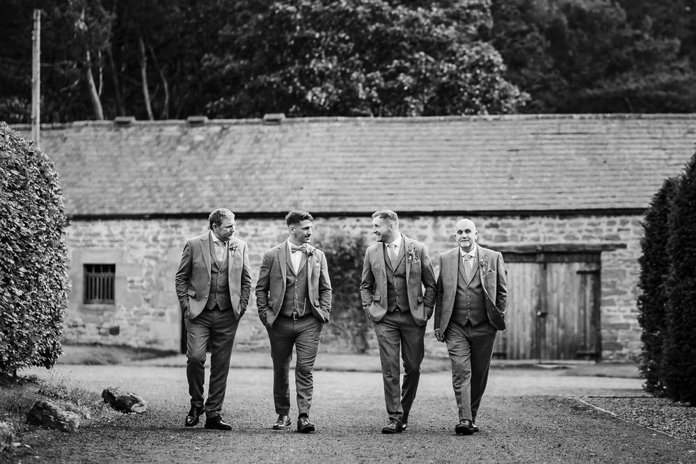 A black and white photo of four groomsmen in matching suits walking together outside a rustic stone building.
