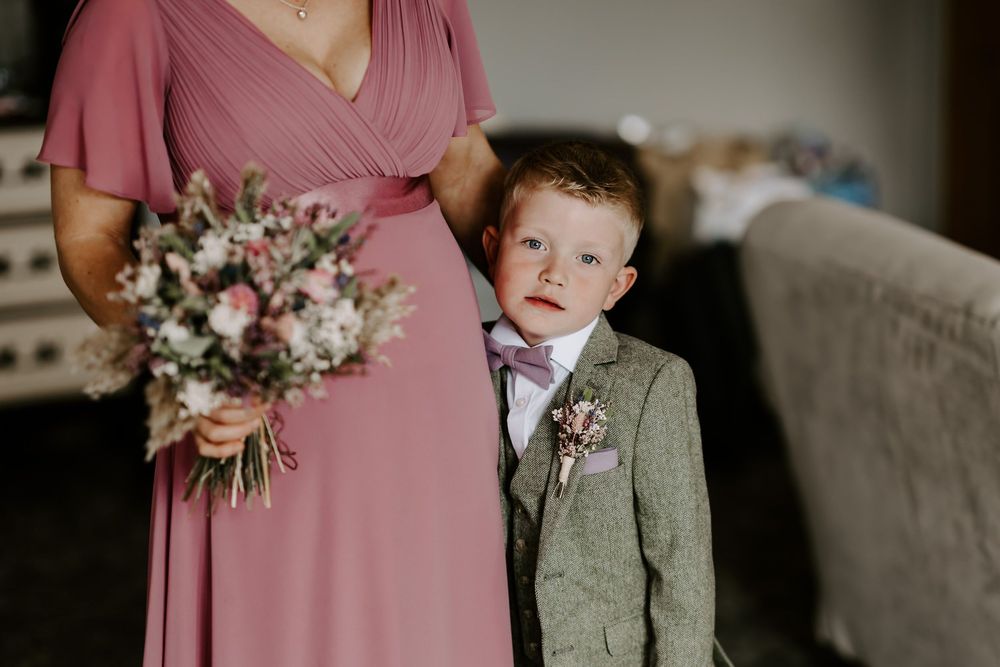 Ring bearer in sage green suit makes funny faces while standing next to pink-dressed bridesmaid holding floral bouquet.