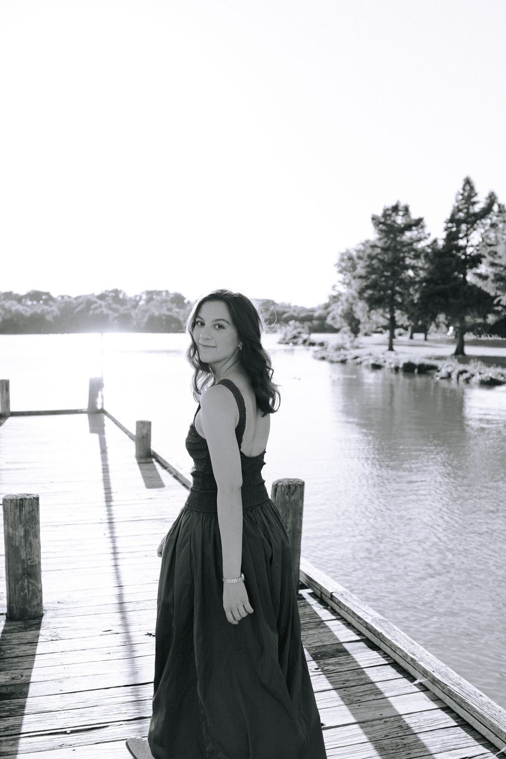 Person wearing a black dress poses artistically on wooden dock over calm waters.