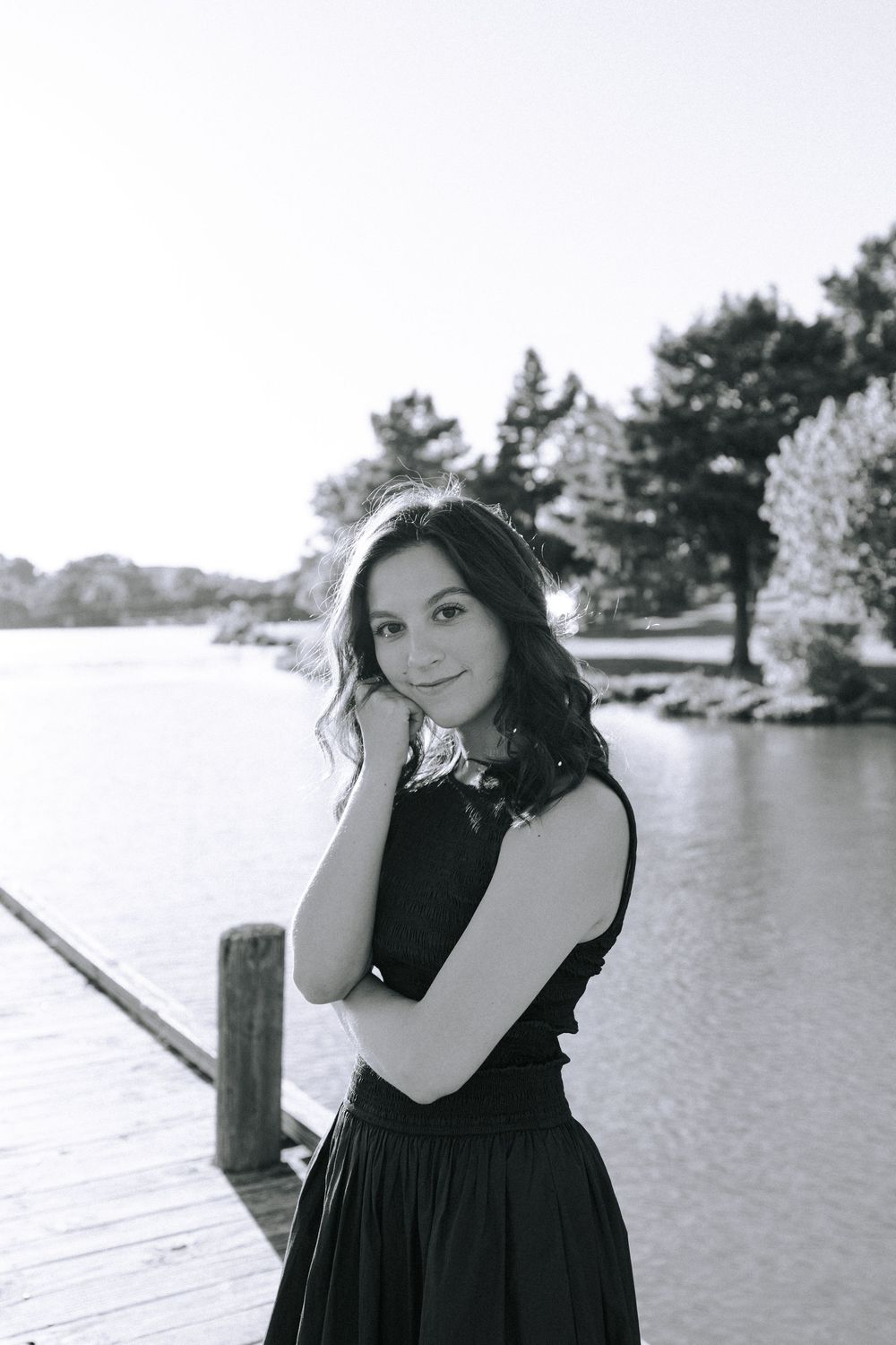 Black and white photo of someone in an elegant dress posing thoughtfully on a dock by the water.