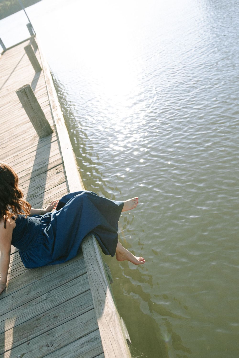 Figure in dark clothing relaxes on edge of wooden pier with feet dangling over water.