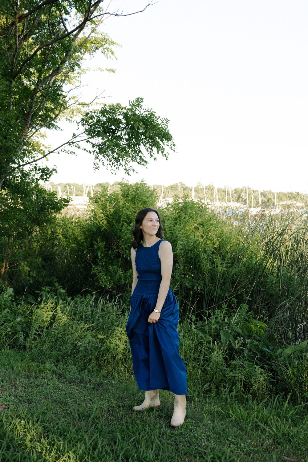 A navy blue maxi dress flows gracefully against a natural backdrop of trees and tall grass at sunset.
