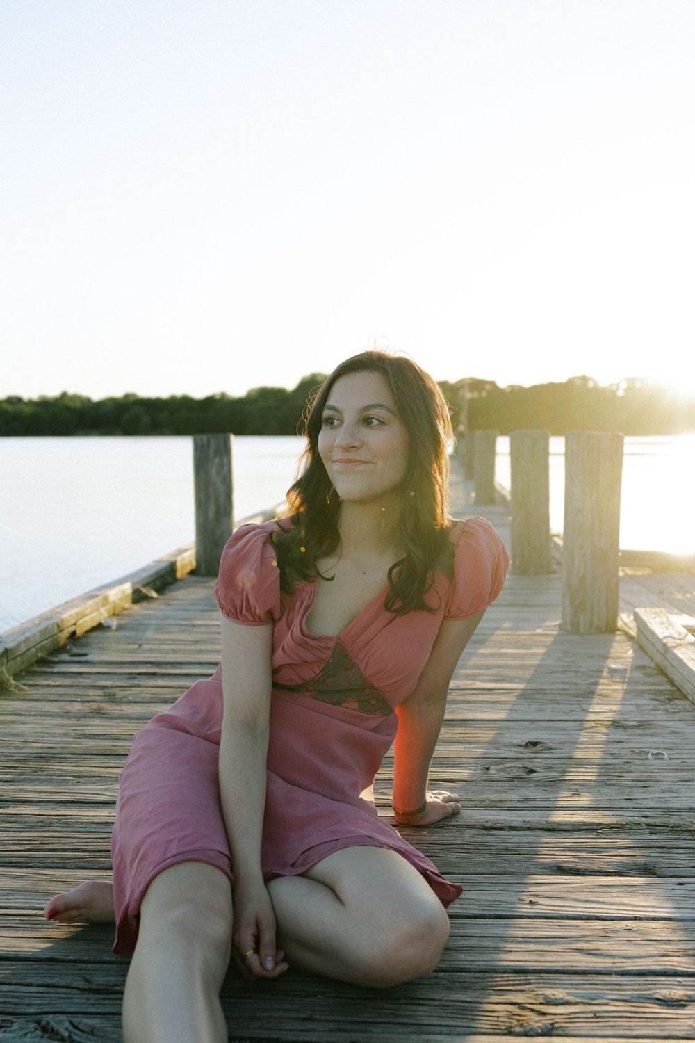 Someone poses gracefully on a wooden pier wearing a pink dress during sunset by the water.