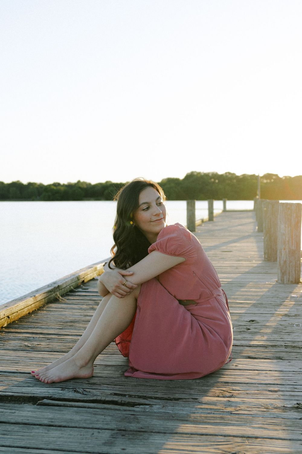 Someone in a pink dress sits contemplatively on a wooden dock during golden hour.