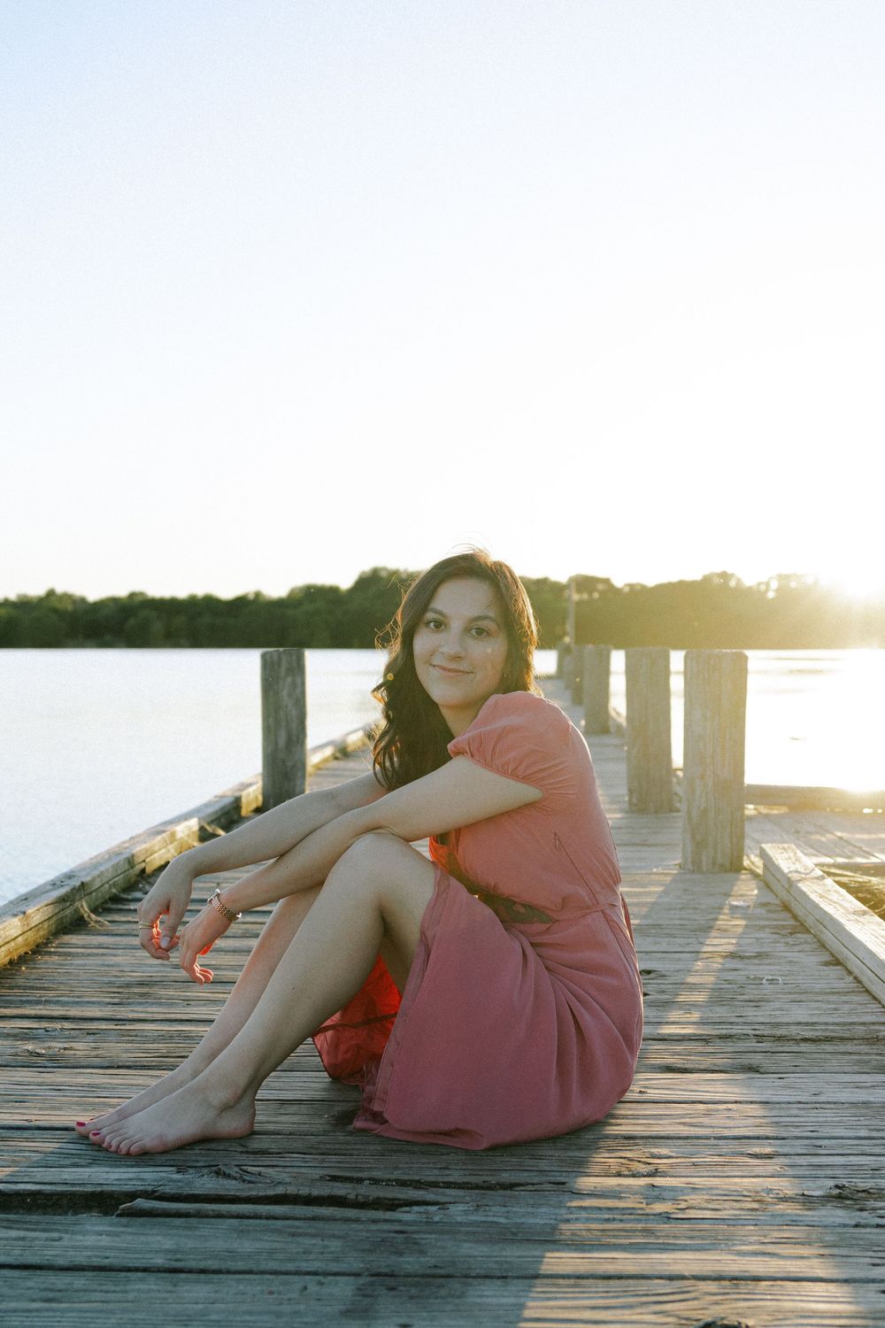 Someone relaxes on a wooden pier wearing a pink dress as the sun sets over calm waters.