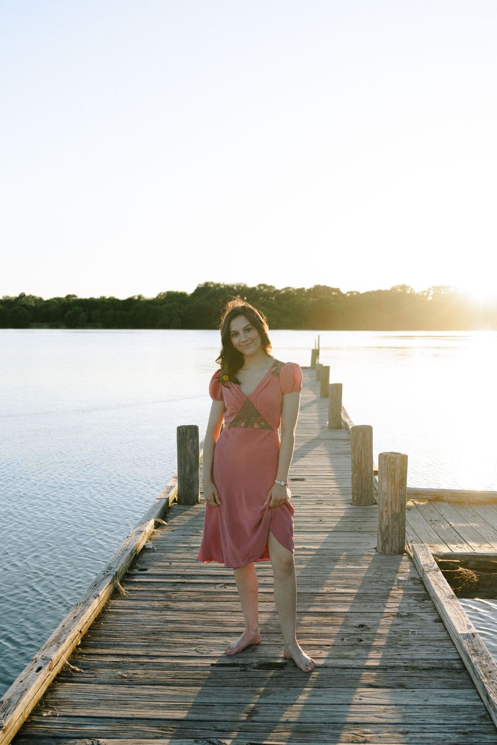 Someone in a pink dress stands on a wooden pier during golden hour.