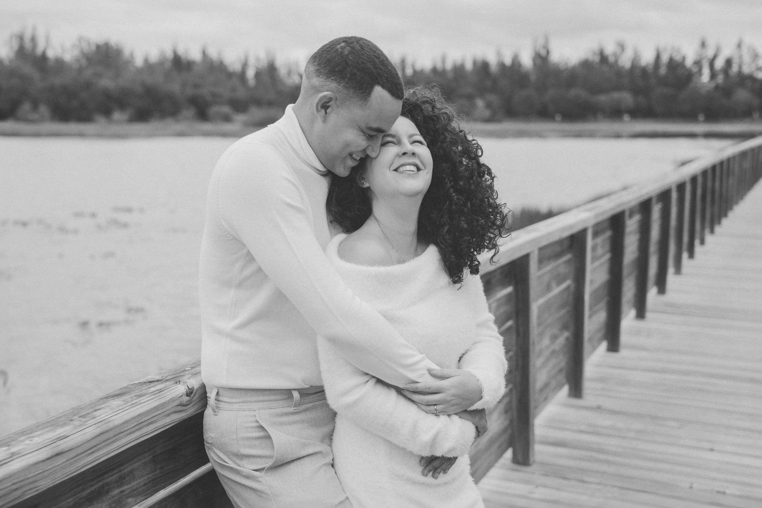 A romantic photoshoot of a couple sharing intimate moments on a wooden boardwalk near the water.