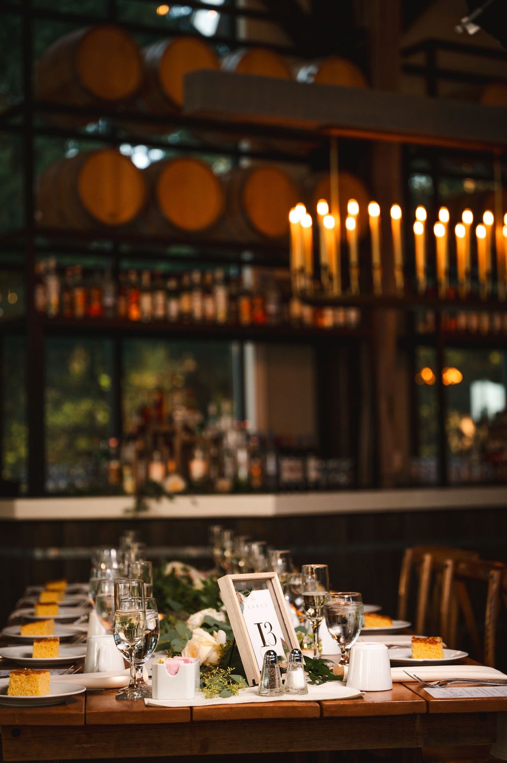 Elegant wedding table setting with glowing candles and wine barrels above bar in background  of reception Barn at Villa Venezia Middletown.