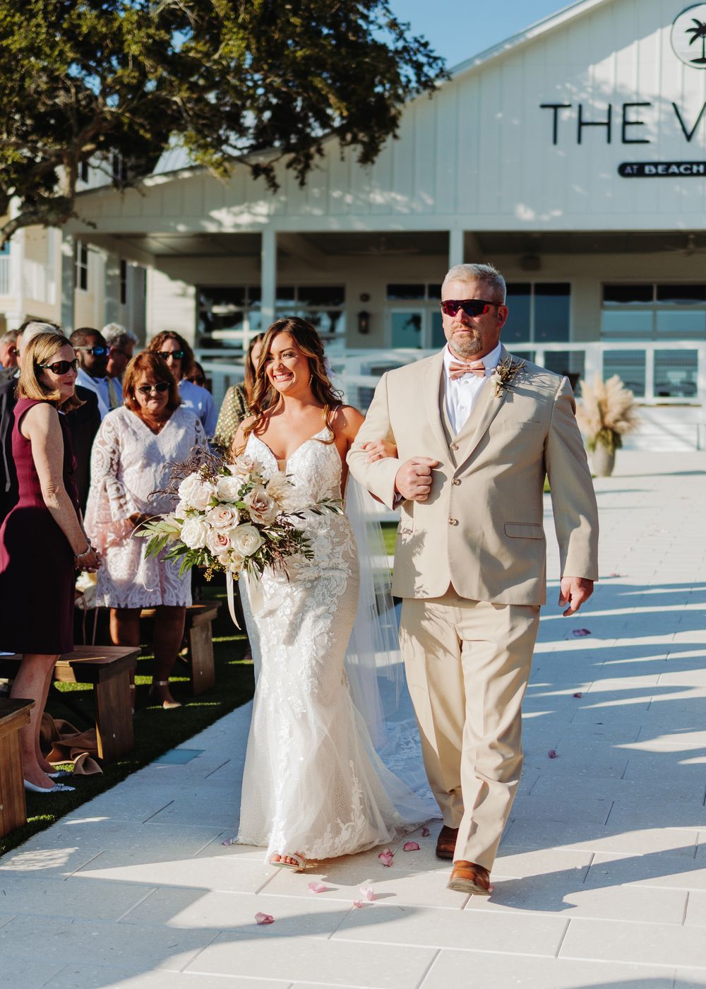 Wedding ceremony processional on white walkway with couple in white and beige formal attire.
